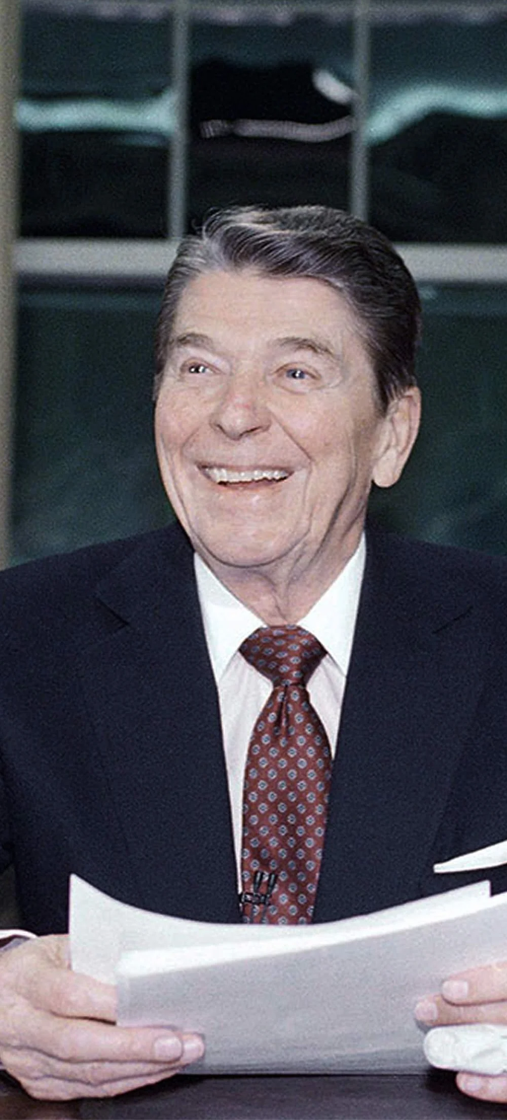 Ronald Reagan, wearing a dark suit, white shirt, and a burgundy patterned tie, is smiling and holding a document. He appears to be in a professional setting.