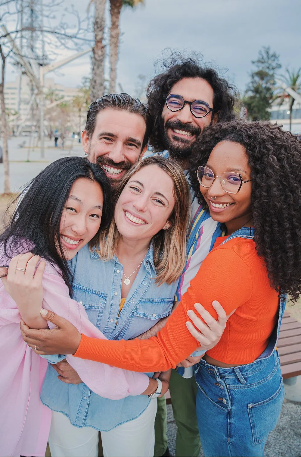 Group of five diverse friends smiling and hugging outdoors on a cloudy day.