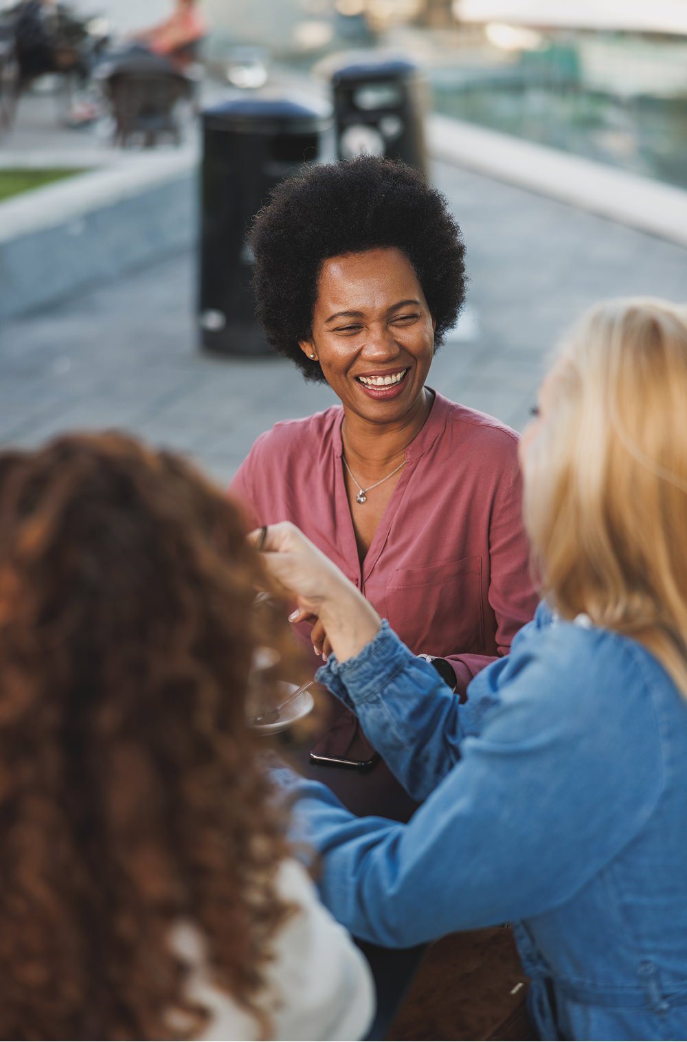 A woman with short curly hair wearing a pink blouse is smiling while talking to two women with blonde hair and red hair, who are sitting at a table outdoors.