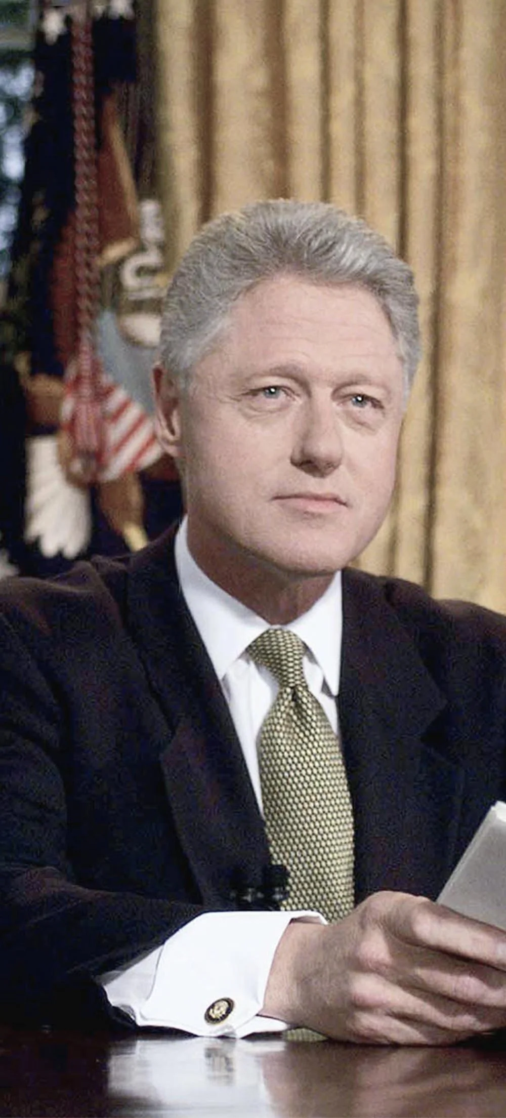 Bill Clinton, wearing a dark suit, white shirt, and yellow patterned tie, sitting at a table in a formal setting, holding a piece of paper.