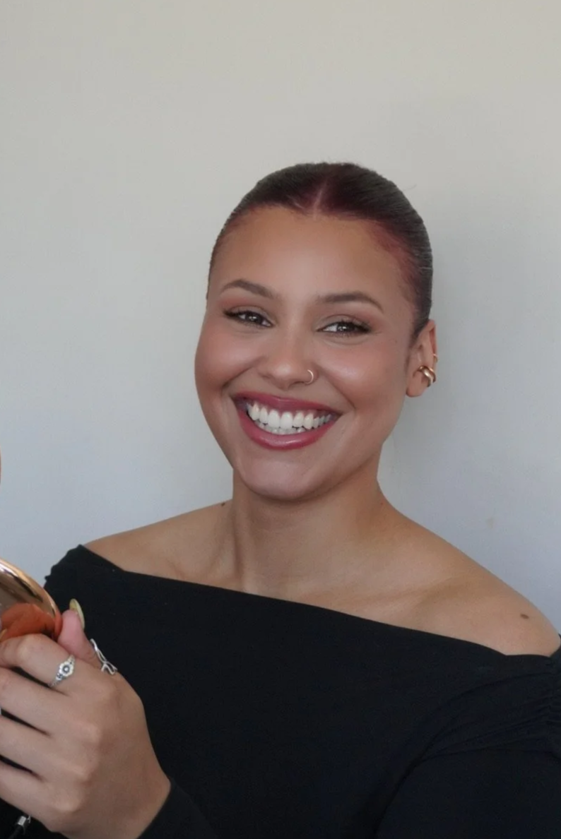 A smiling woman with dark hair slicked back, holding a phone, wearing earrings and a black off-shoulder top, standing against a plain light-colored wall.