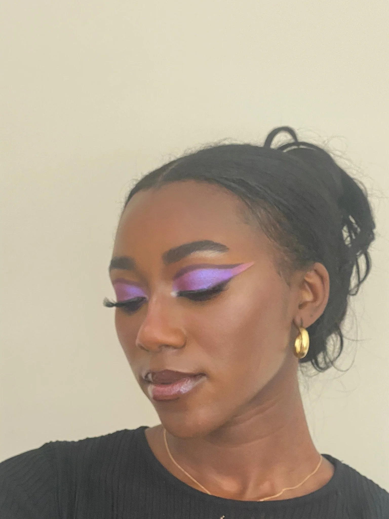 A woman with flawless skin and dramatic purple and pink eye makeup, wearing gold earrings, a gold necklace, and a black top, elegant hairstyle, and closed eyes.