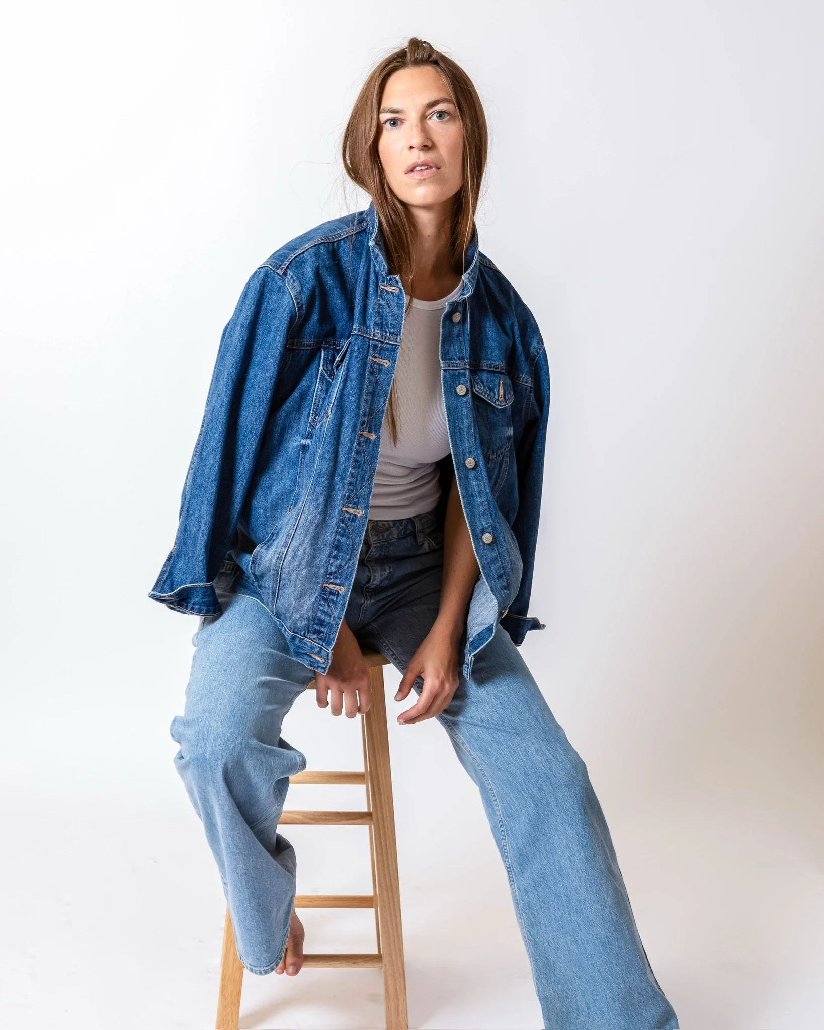A woman with long brown hair sitting on a wooden stool against a plain white background, wearing a blue denim jacket, white T-shirt, and loose-fitting jeans.