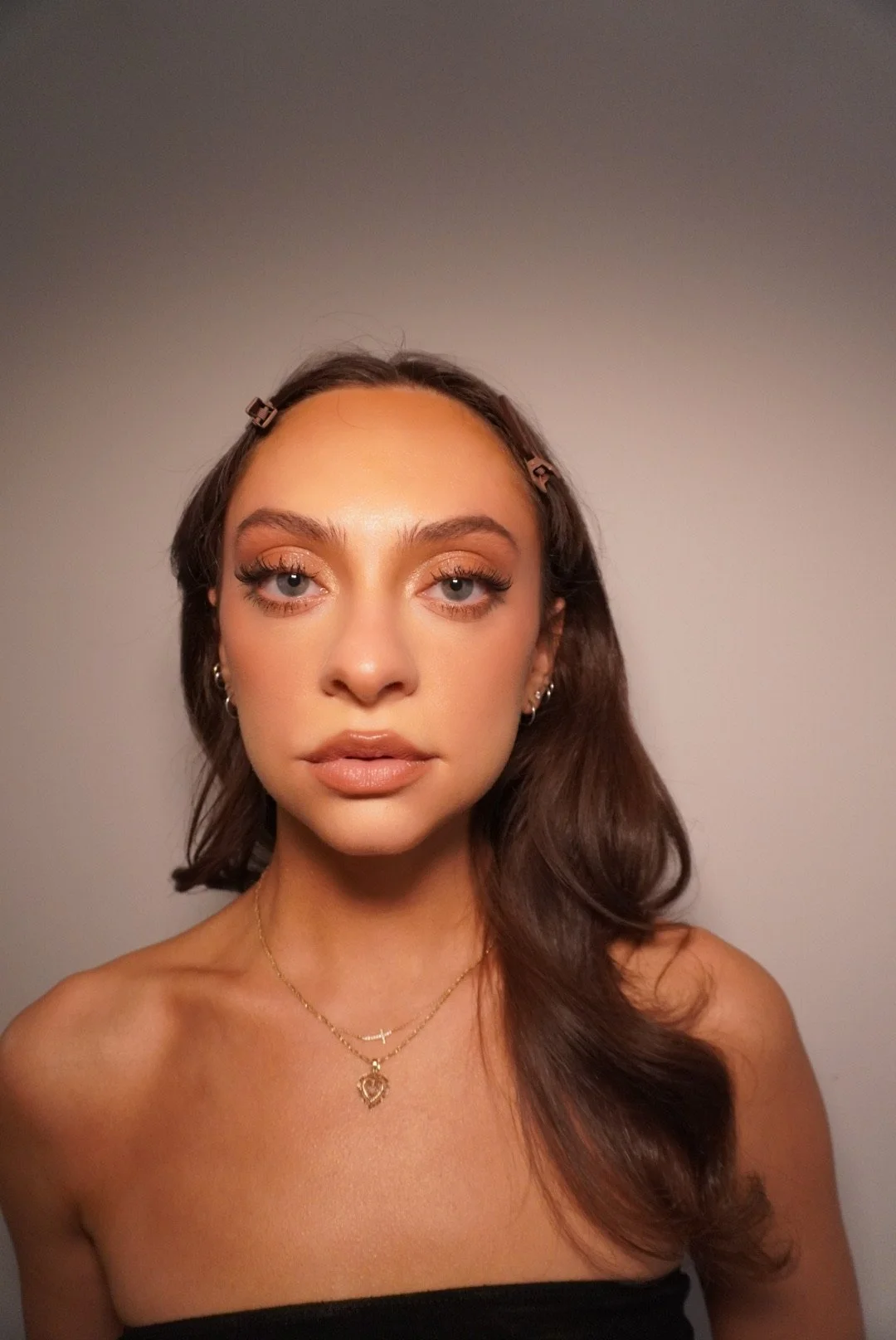A young woman with shoulder-length brown hair, styled with small hair clips, wearing makeup with gold eyeshadow and nude lipstick, gold earrings, and layered gold necklaces, against a plain, neutral background.