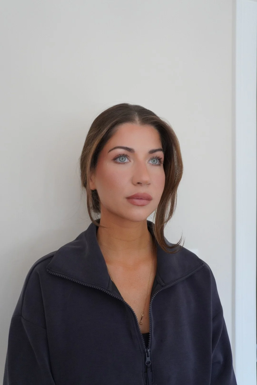 A woman with brown hair and blue eyes sitting against a white wall, wearing a dark zip-up jacket.