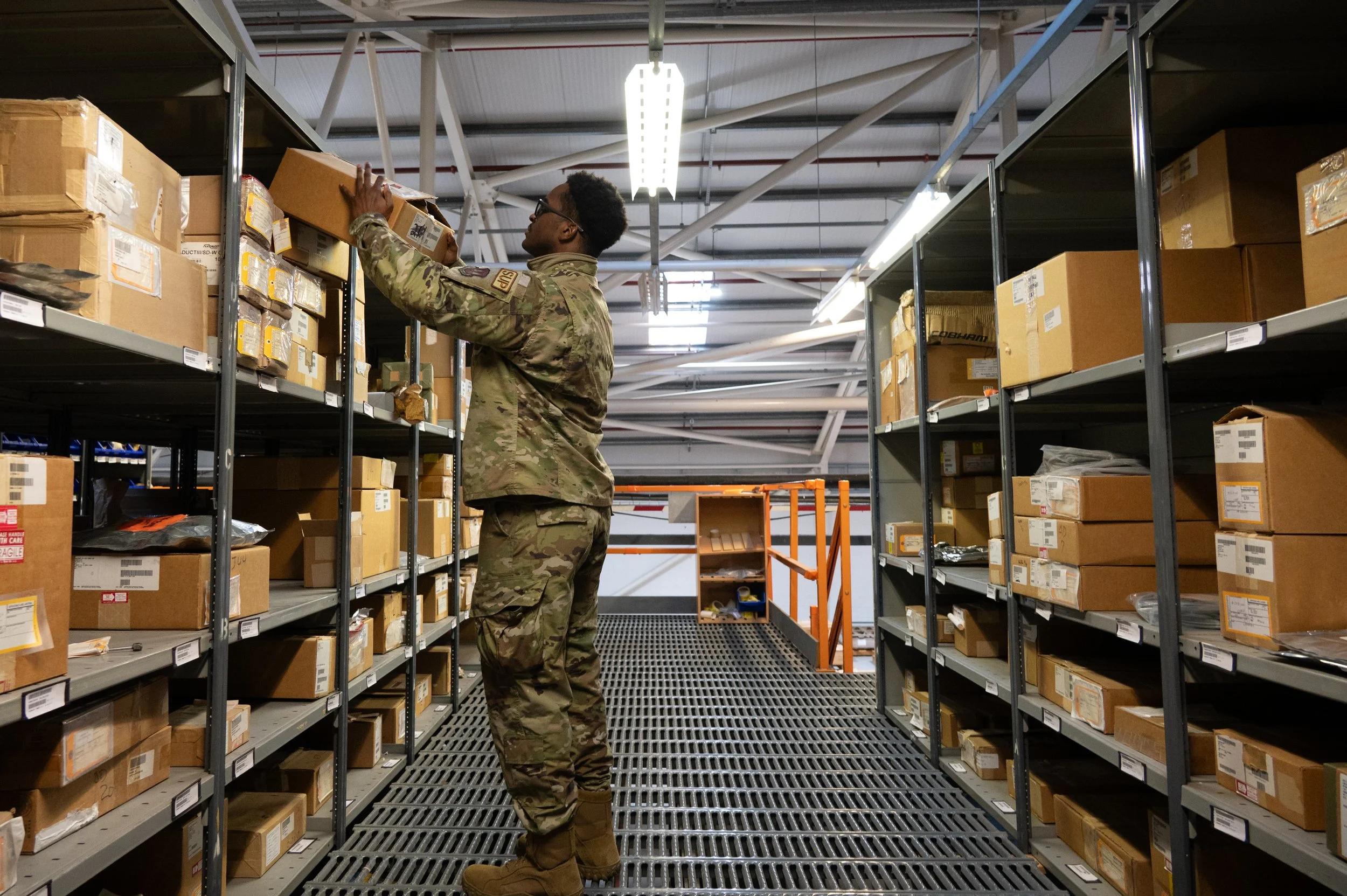 A person in military camouflage uniform stocking shelves with boxes in a warehouse.