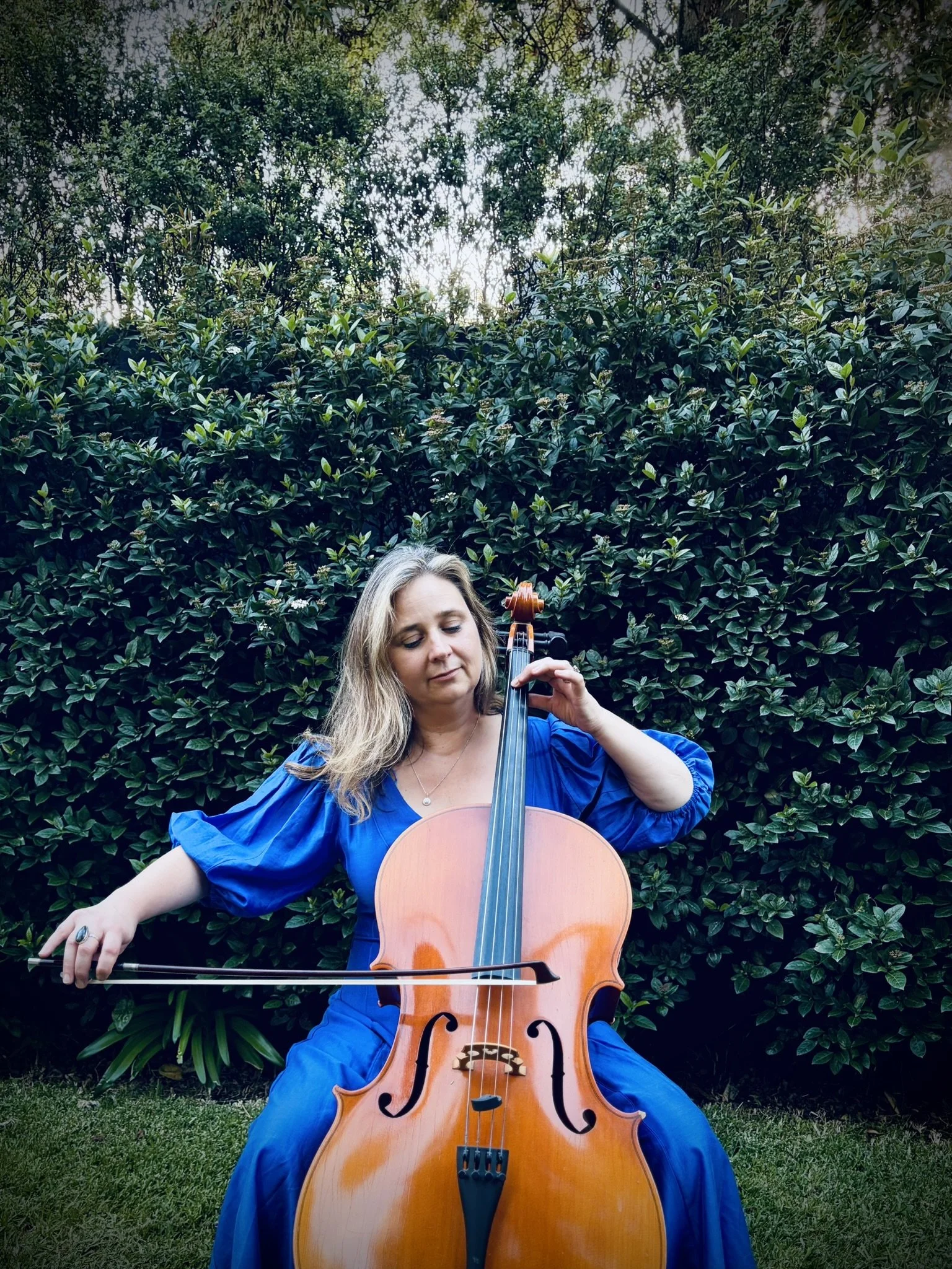 A woman with long blond hair wearing a vibrant blue dress playing a cello outdoors in front of lush green foliage.