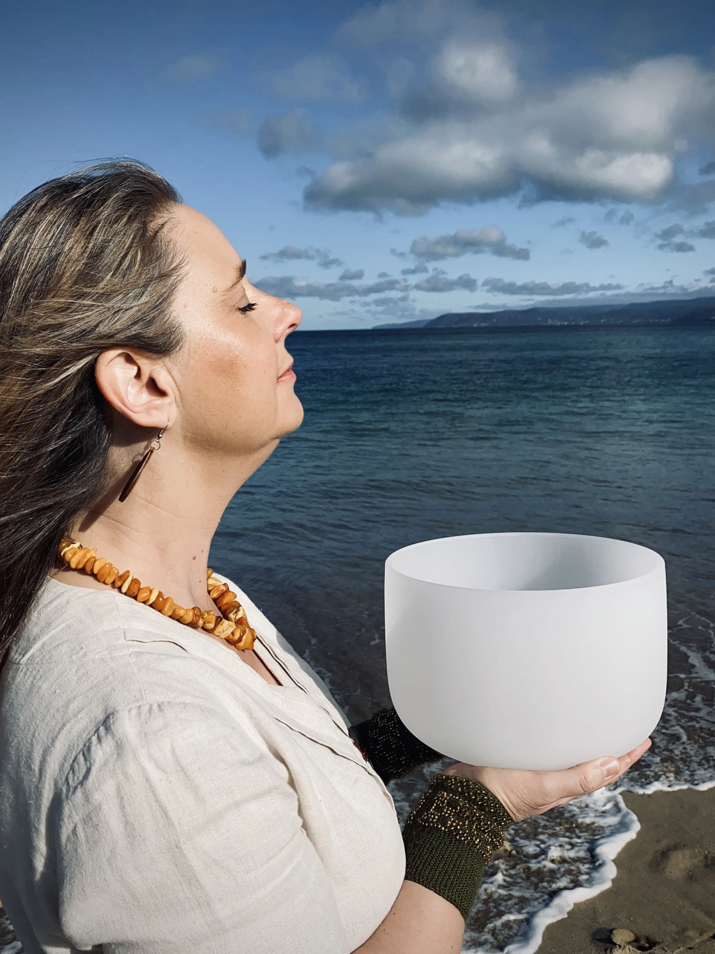 A woman with long hair and beaded jewelry is standing by the shore holding a large white singing bowl. She has her eyes closed and appears to be meditating or enjoying the peaceful environment. The ocean, sky with clouds, and distant land are visible in the background.