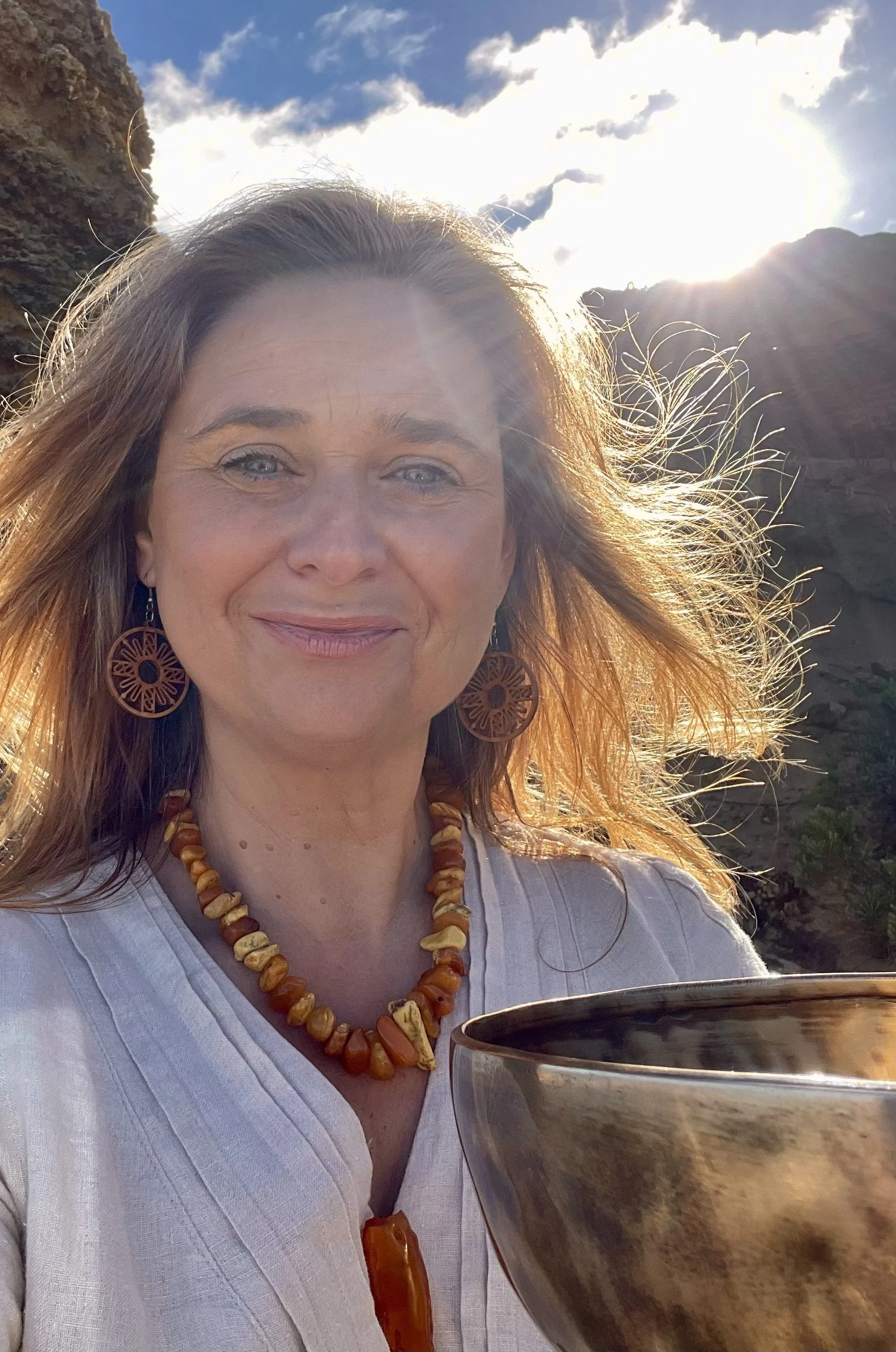 Woman with long, light brown hair, wearing large wooden earrings and an amber beaded necklace, smiling while holding a Himalayan bowl outdoors on a sunny day with a rocky background.
