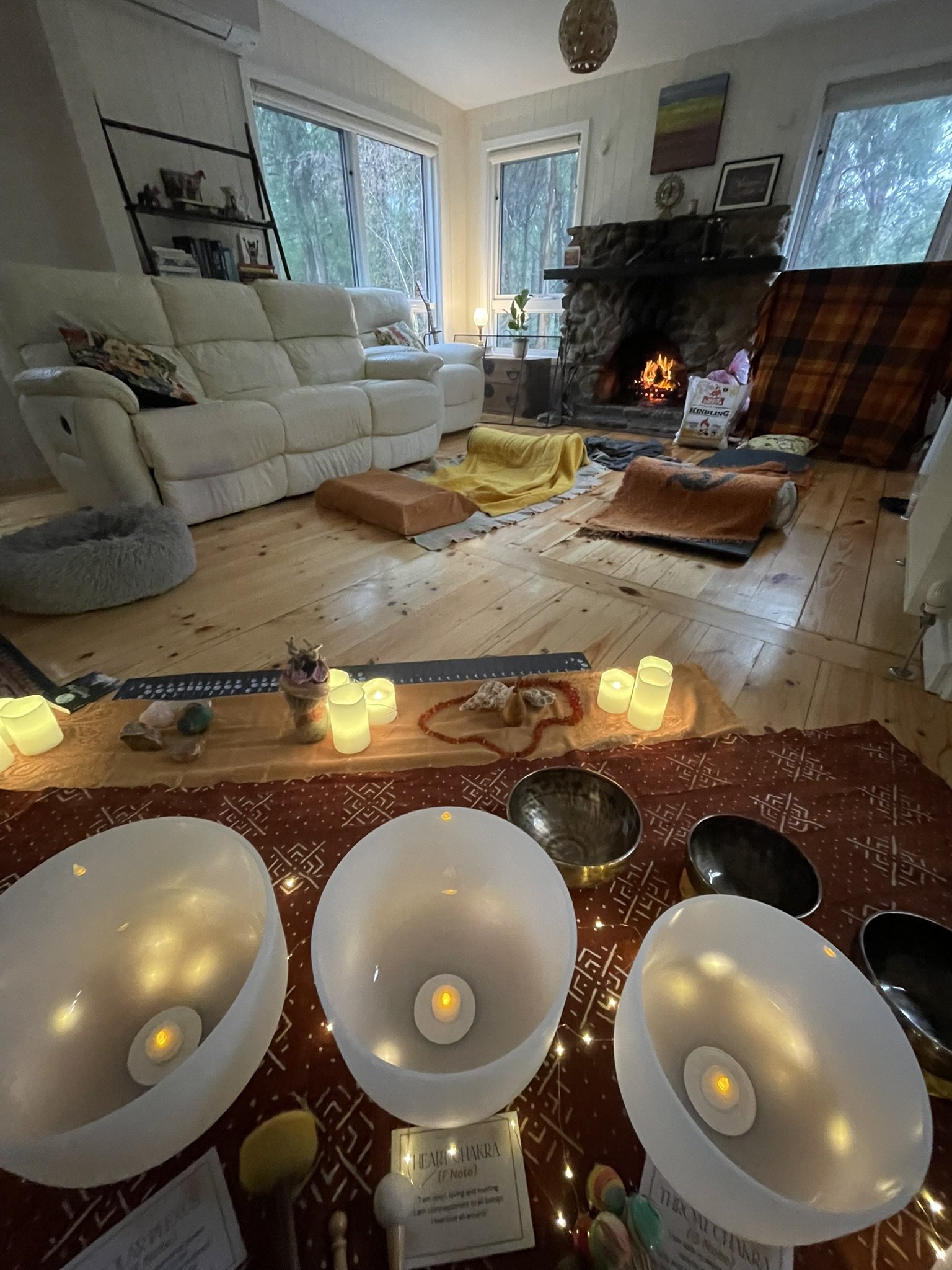 A cozy living room with a white couch, a stone fireplace with a small fire, and large windows showing trees outside. The foreground shows a table with candles and decorative items, and the room has a warm, inviting ambiance.
