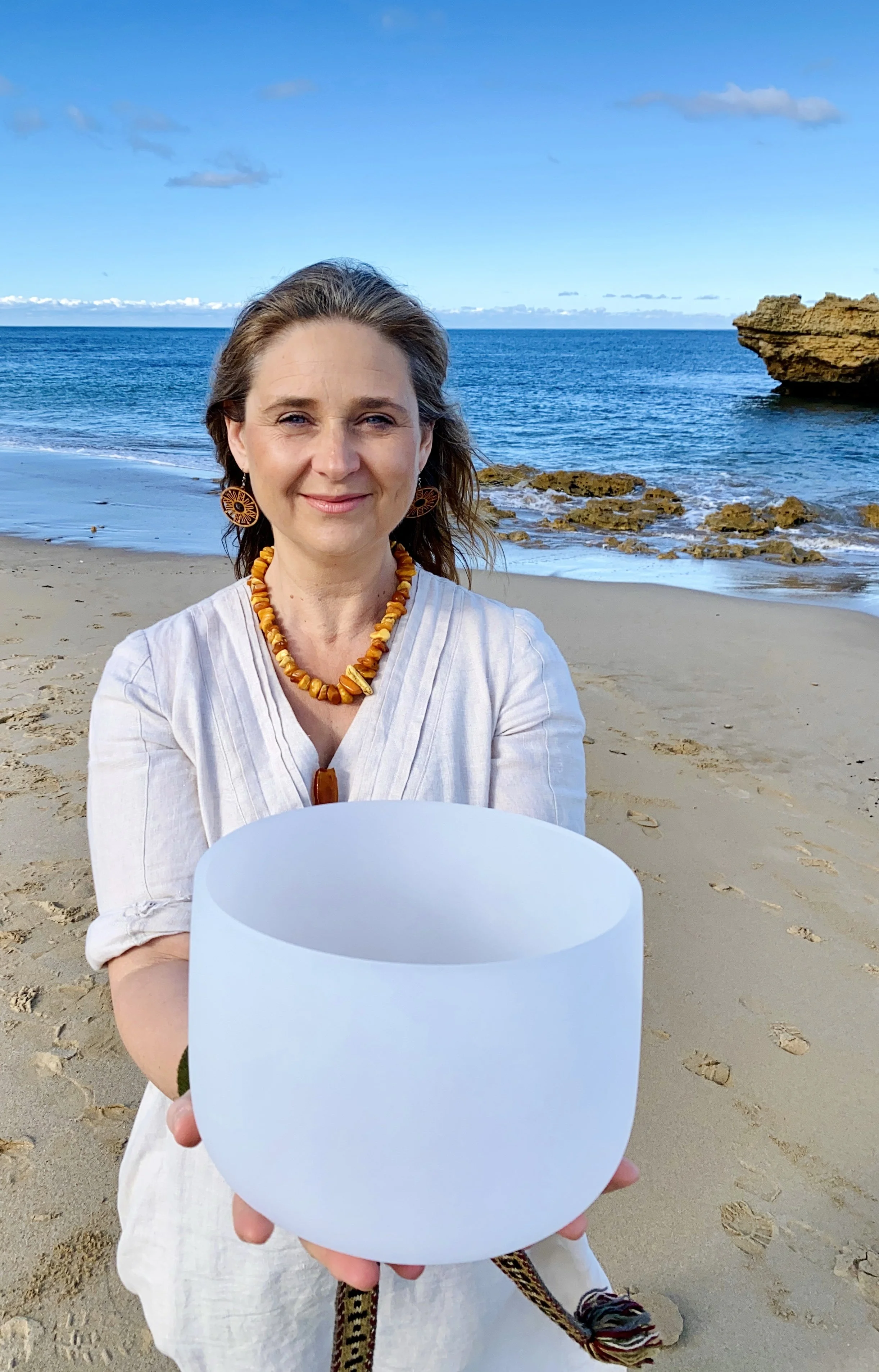 A woman standing on a sandy beach holding a crystal bowl. She has fair hair, is wearing light linen clothing, amber beaded jewelry, and wooden earrings. The ocean, rocks, and a blue sky with some clouds are visible in the background.