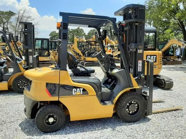 A yellow CAT forklift with black accents parked on a gravel lot, surrounded by other construction vehicles and equipment, under a partly cloudy sky.