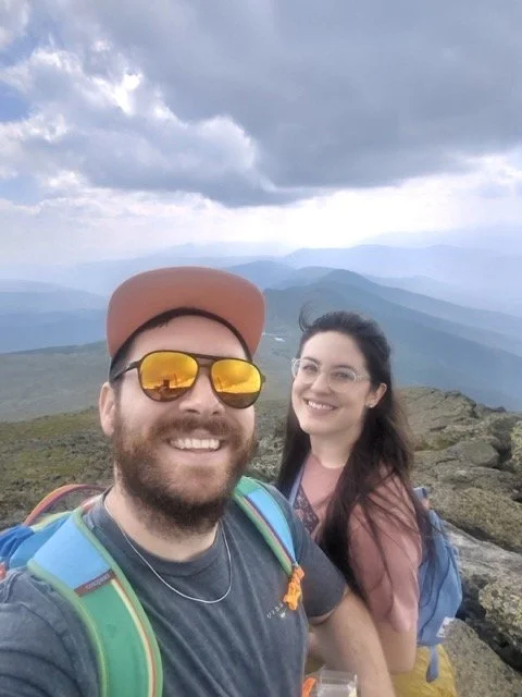 Two smiling hikers, a man with sunglasses and a woman, taking a selfie on a mountain trail with a cloudy mountain landscape in the background.
