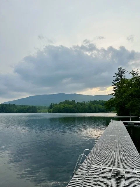A dock extending over a calm lake with a mountain and cloudy sky in the background, surrounded by trees.