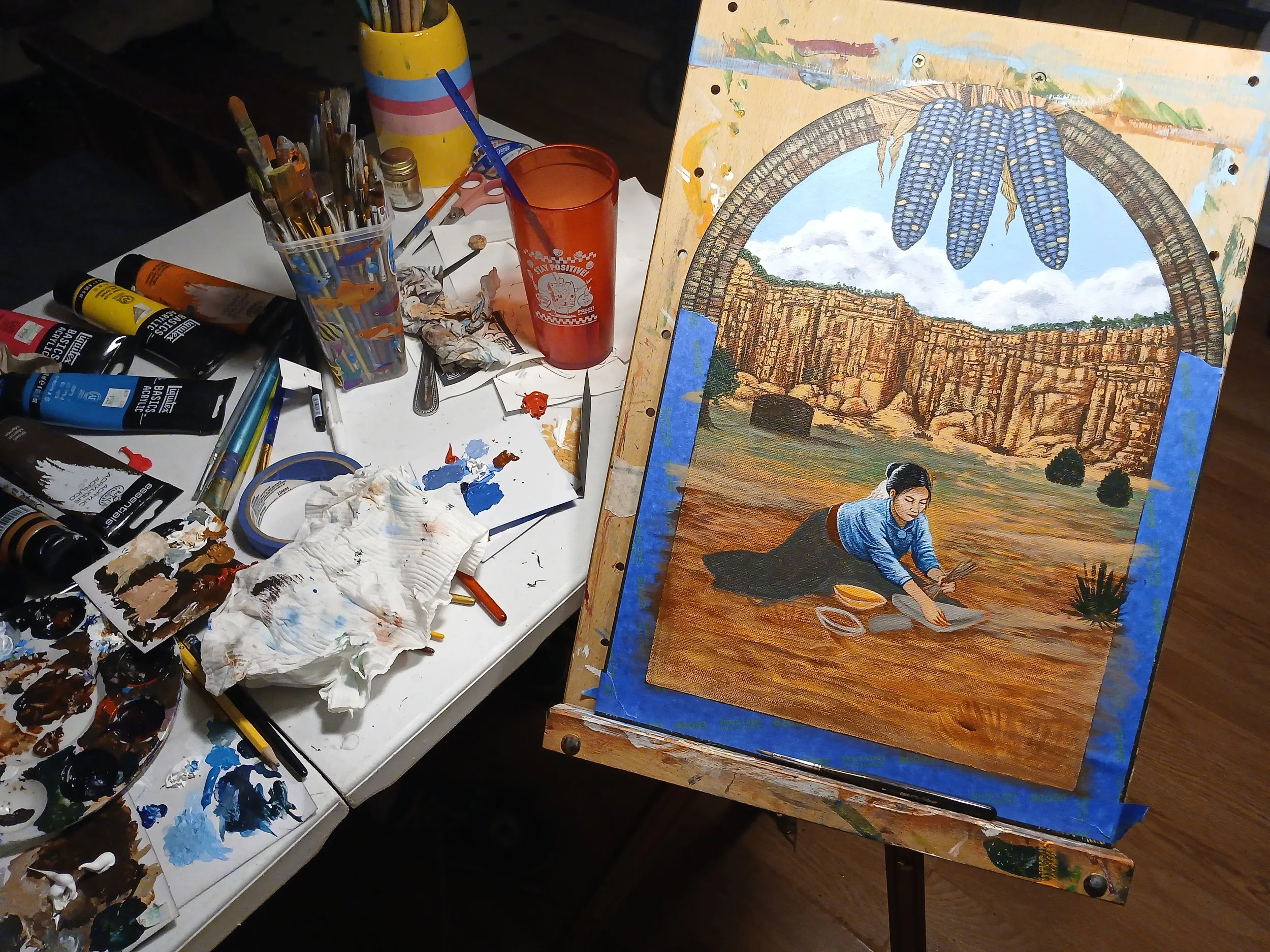 A work in progress shot of a painting featuring a young woman grinding blue corn in front of a wall of red rocks as a roadrunner flits across. Sold during the 2025 Totah Festival.