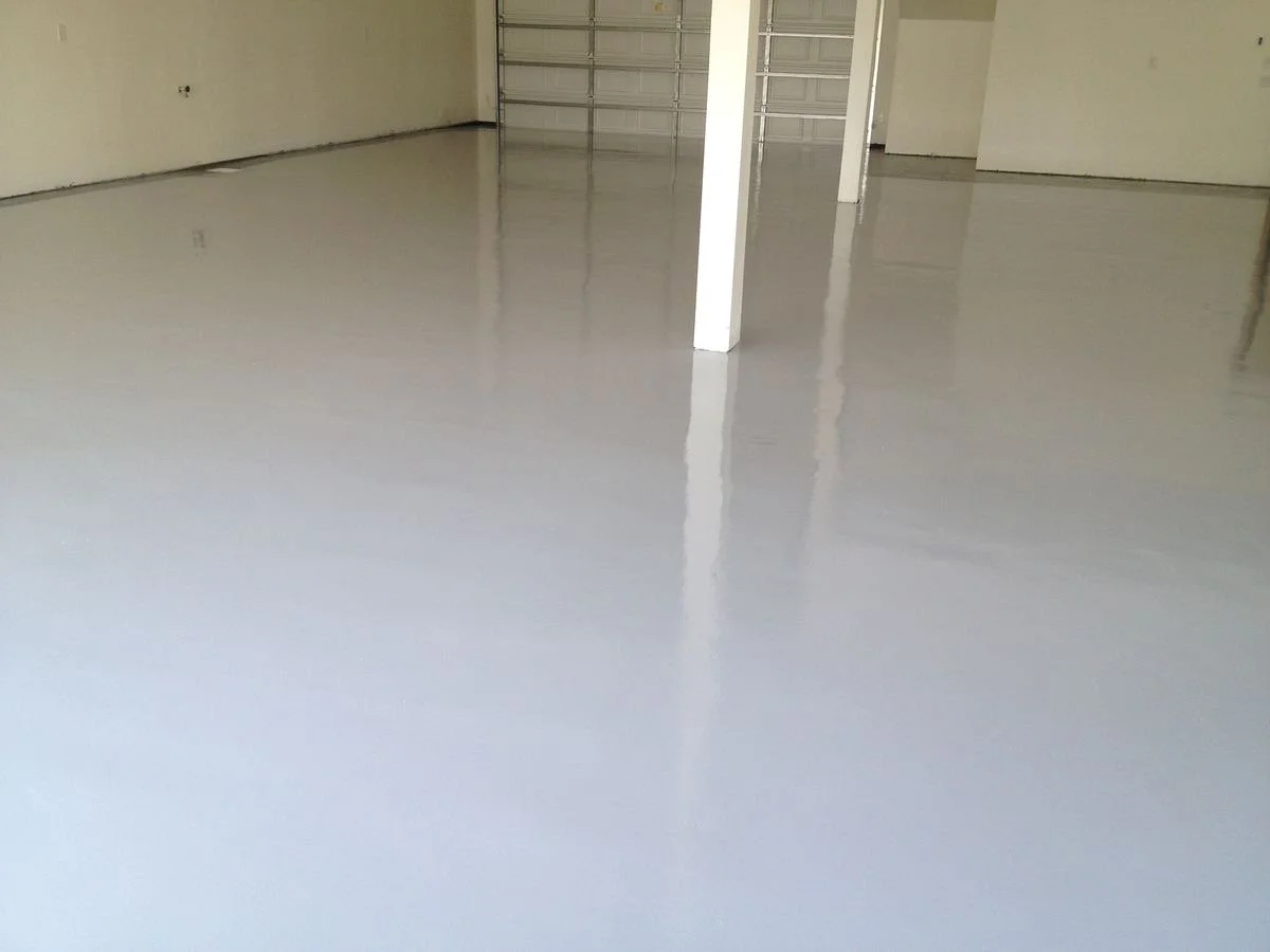 Interior of an unfinished garage with a shiny epoxy coated concrete floor, walls, and a closed garage door in the background.