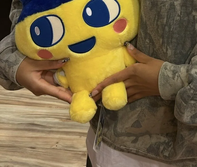 Person holding a large tamagotchi plush toy with a happy cartoon face, blue eyes, and red cheeks.