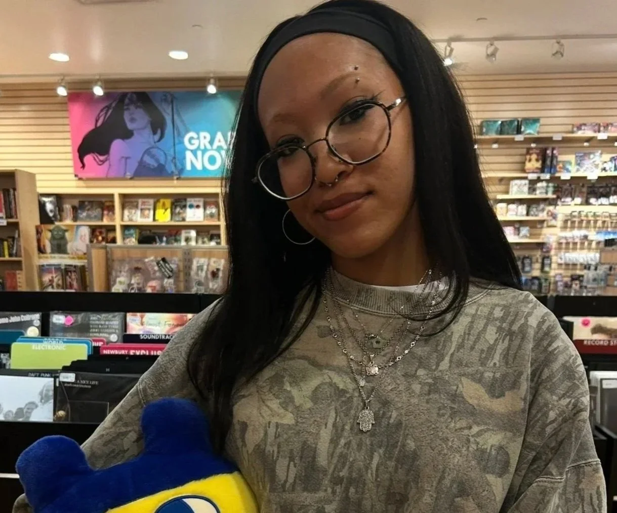 Young woman with bleached brows, long black hair, glasses, and layered necklaces standing inside a record store, holding a blue and yellow stuffed toy. Wearing real-tree camo, or camo sweatshirt. Holding a tamagotchi plush.