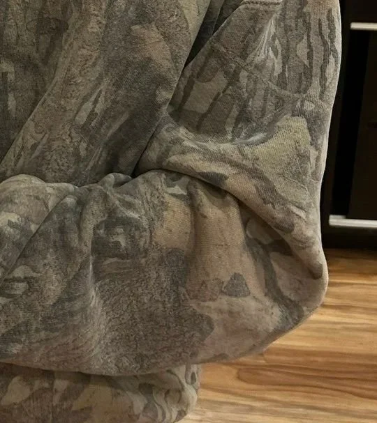 Close-up of a real-tree camouflage-patterned fabric, possibly a sweatshirt or hoodie, with a wooden floor in the background.