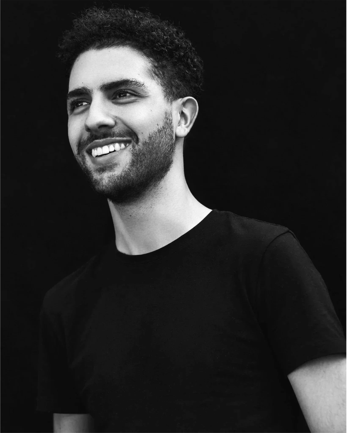 Black-and-white portrait of a smiling young man with curly hair, beard, and wearing a black T-shirt, against a black background.