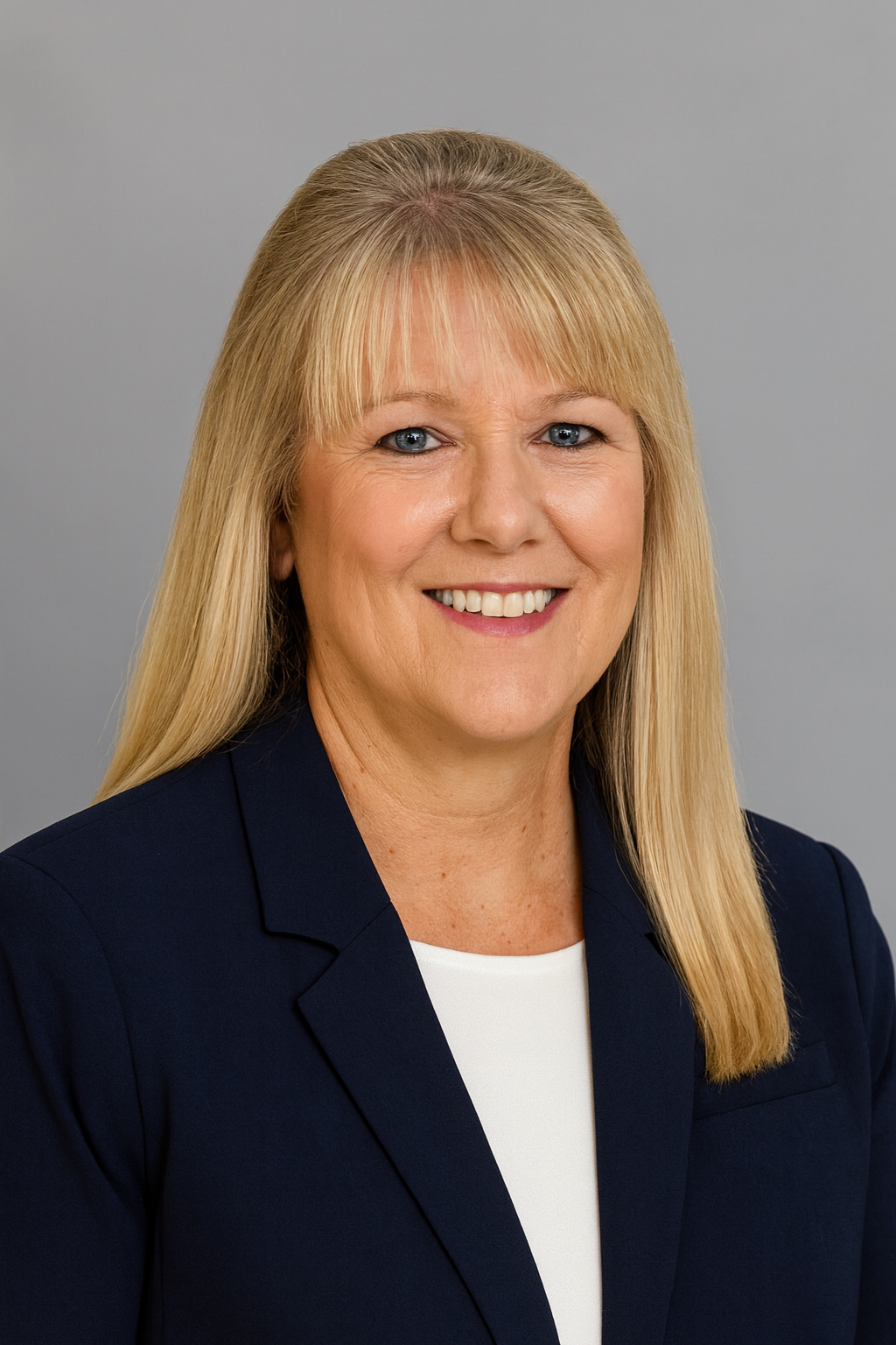 A professional portrait of a smiling woman with long blonde hair, wearing a navy blazer and white top, against a plain gray background.