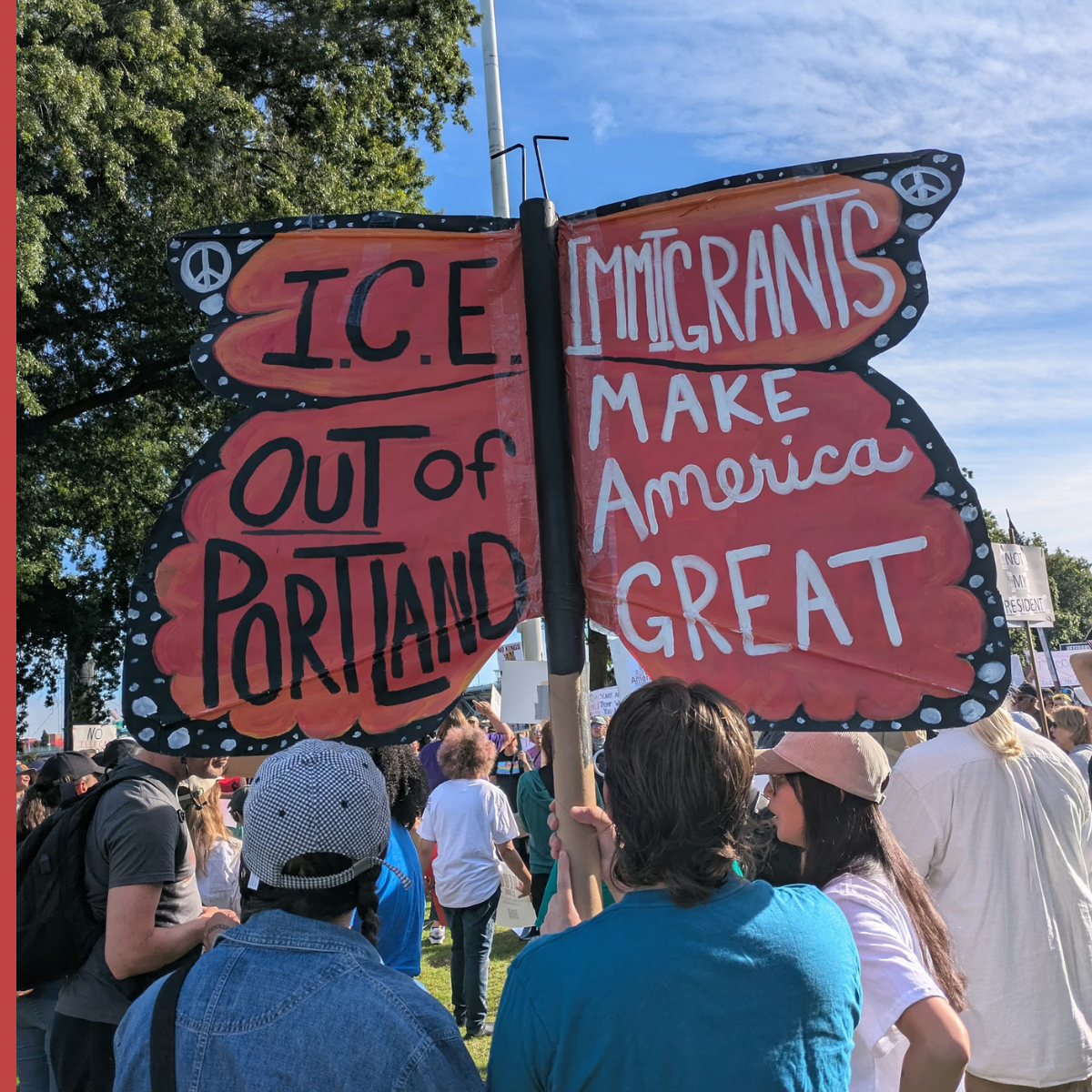 A protest sign at a demonstration with a crowd of people in the background. The sign has a peace symbol on the top left and right corners, with the left side reading, "I.C.E OUT of PORTLAND" and the right side saying, "IMMIGRANTS MAKE America GREAT."