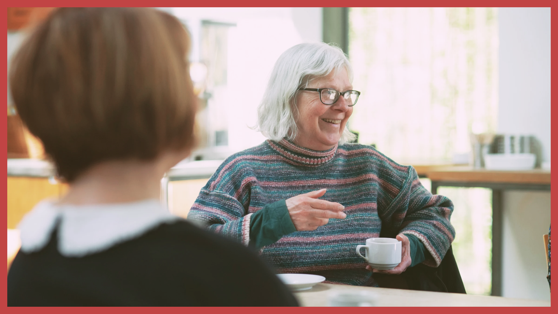 An elderly woman with gray hair and glasses wearing a multicolored striped sweater, smiling and holding a white mug, engaging in conversation with a person with reddish-brown hair, in a bright, cozy kitchen or dining area.