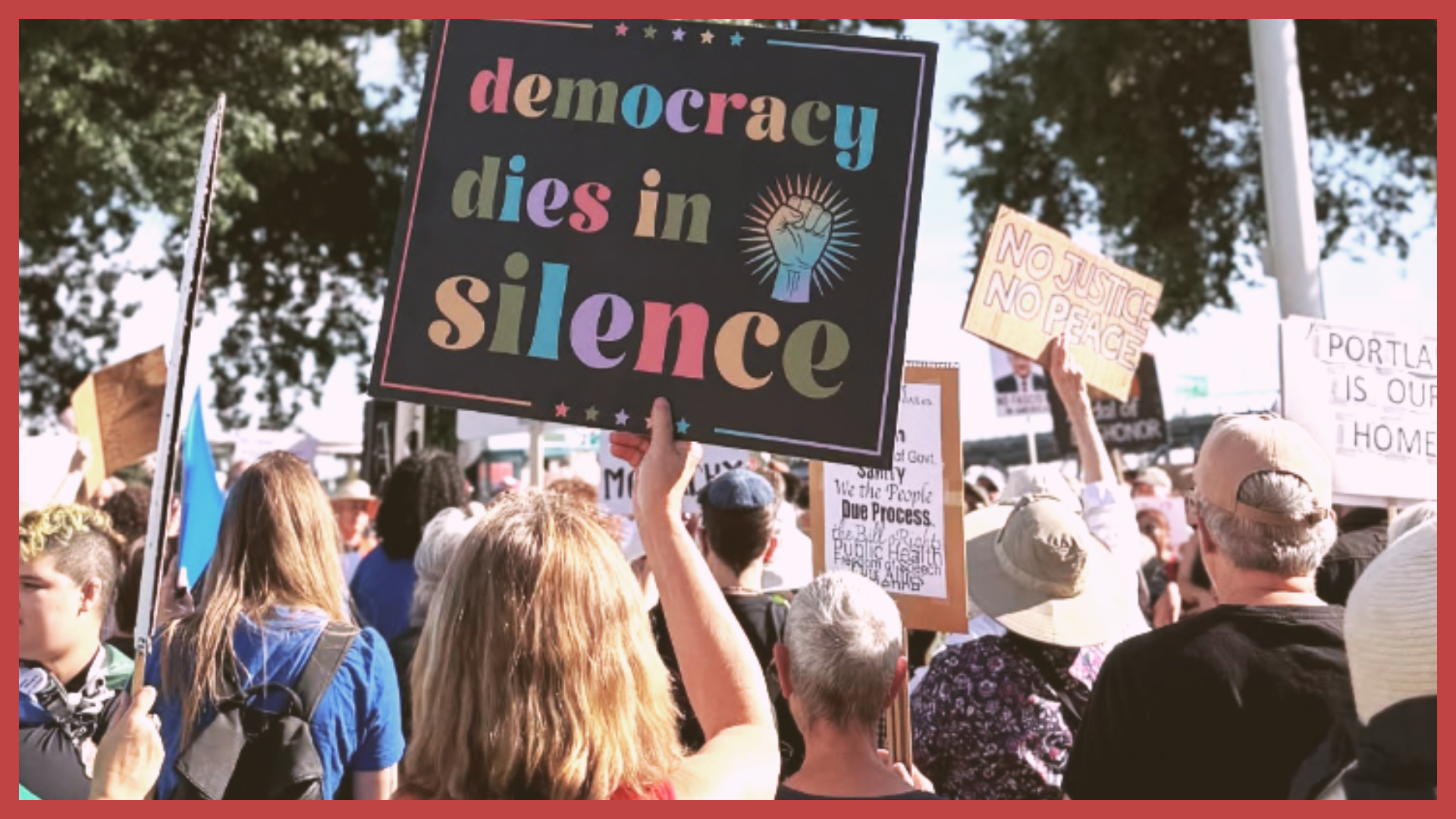 Crowd of people at a protest holding signs, with a prominent sign reading 'democracy dies in silence' and various other signs supporting justice and peace.