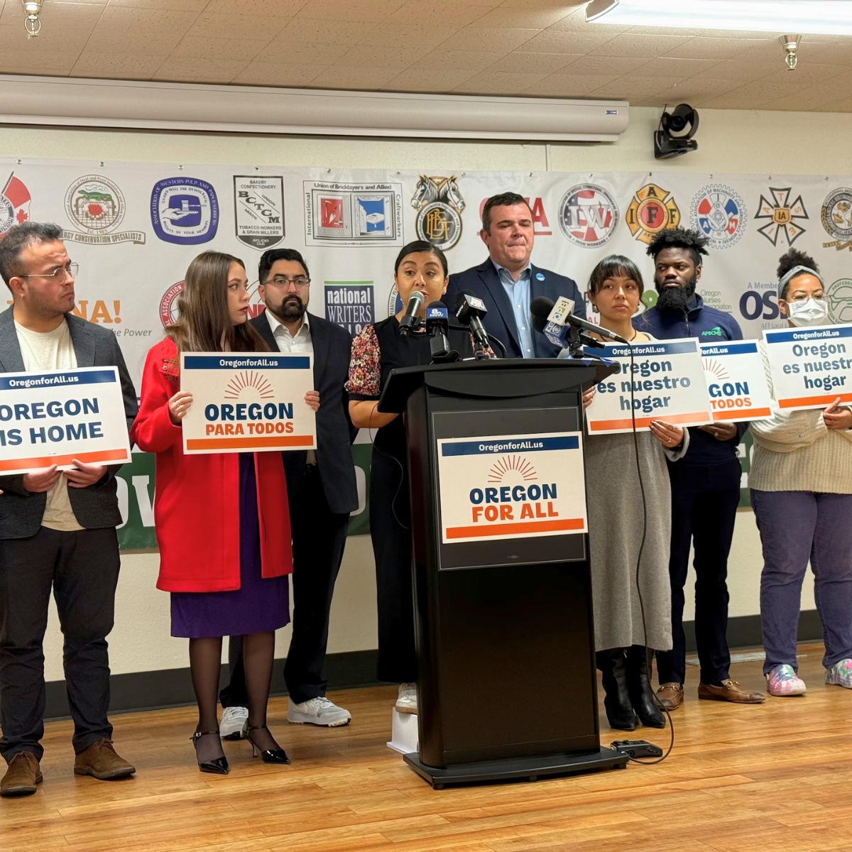 A group of diverse people standing behind a podium, holding signs with messages like "Oregon para todos" and "Oregon is our home," during a press conference