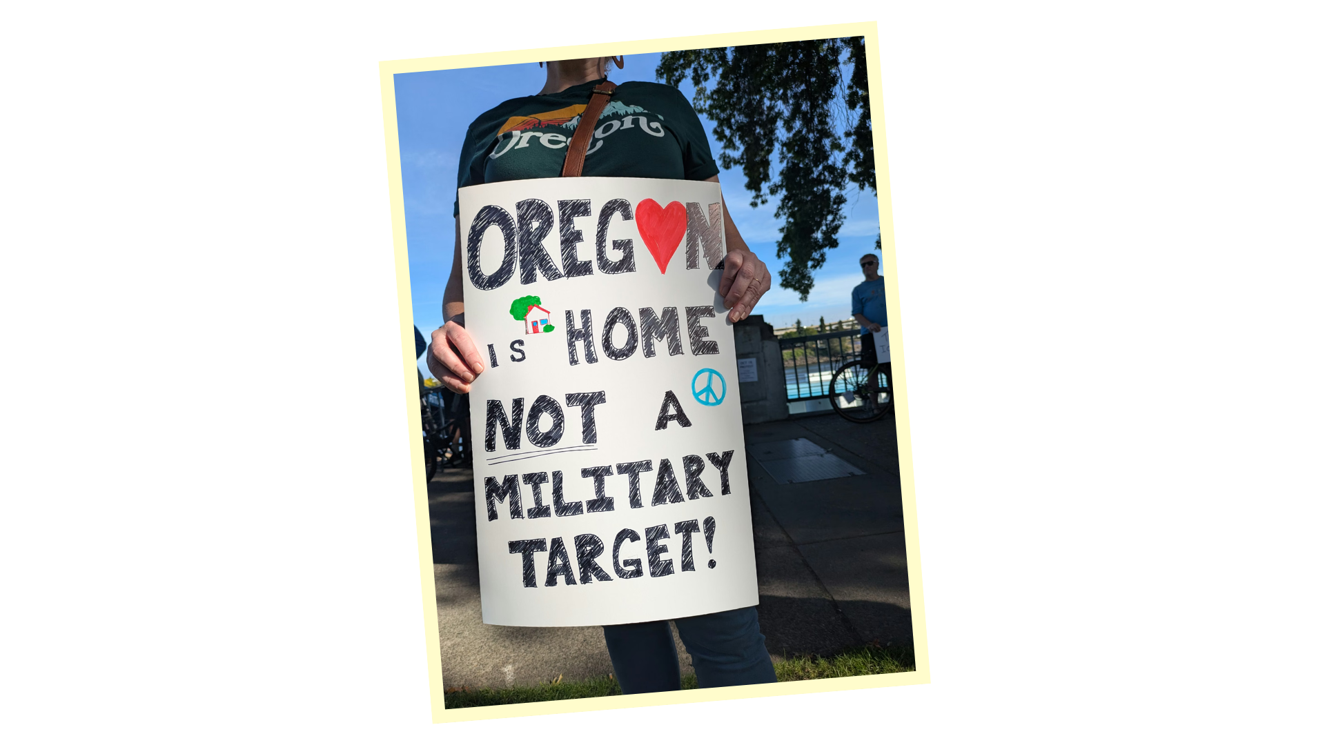 Protestor holding a sign that says "Oregon is Home, Not a Military Target" with a peace symbol, a house, and a heart illustration, at an outdoor demonstration near a body of water and a bike, with a person wearing sunglasses in the background.