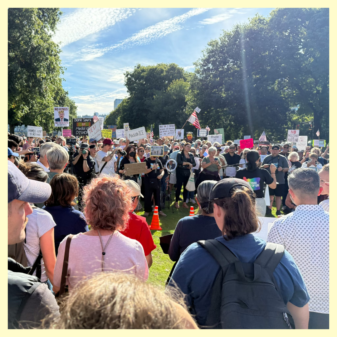 Crowd of people gathered outdoors holding various signs and flags, protesting or rallying on a sunny day with trees and a blue sky in the background.