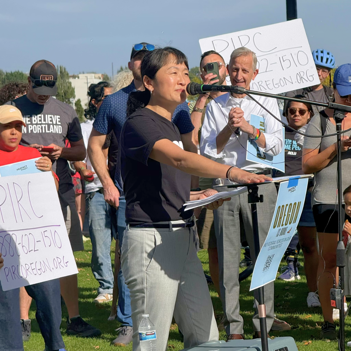 A woman speaking at a rally or protest with a microphone, surrounded by people holding sign