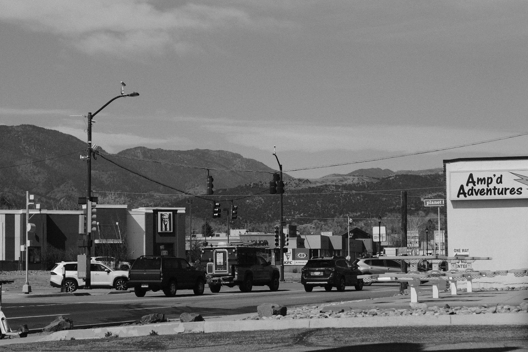 Black and white photo of a small parking lot and street with vehicles, traffic lights, and signs, with mountains in the background.