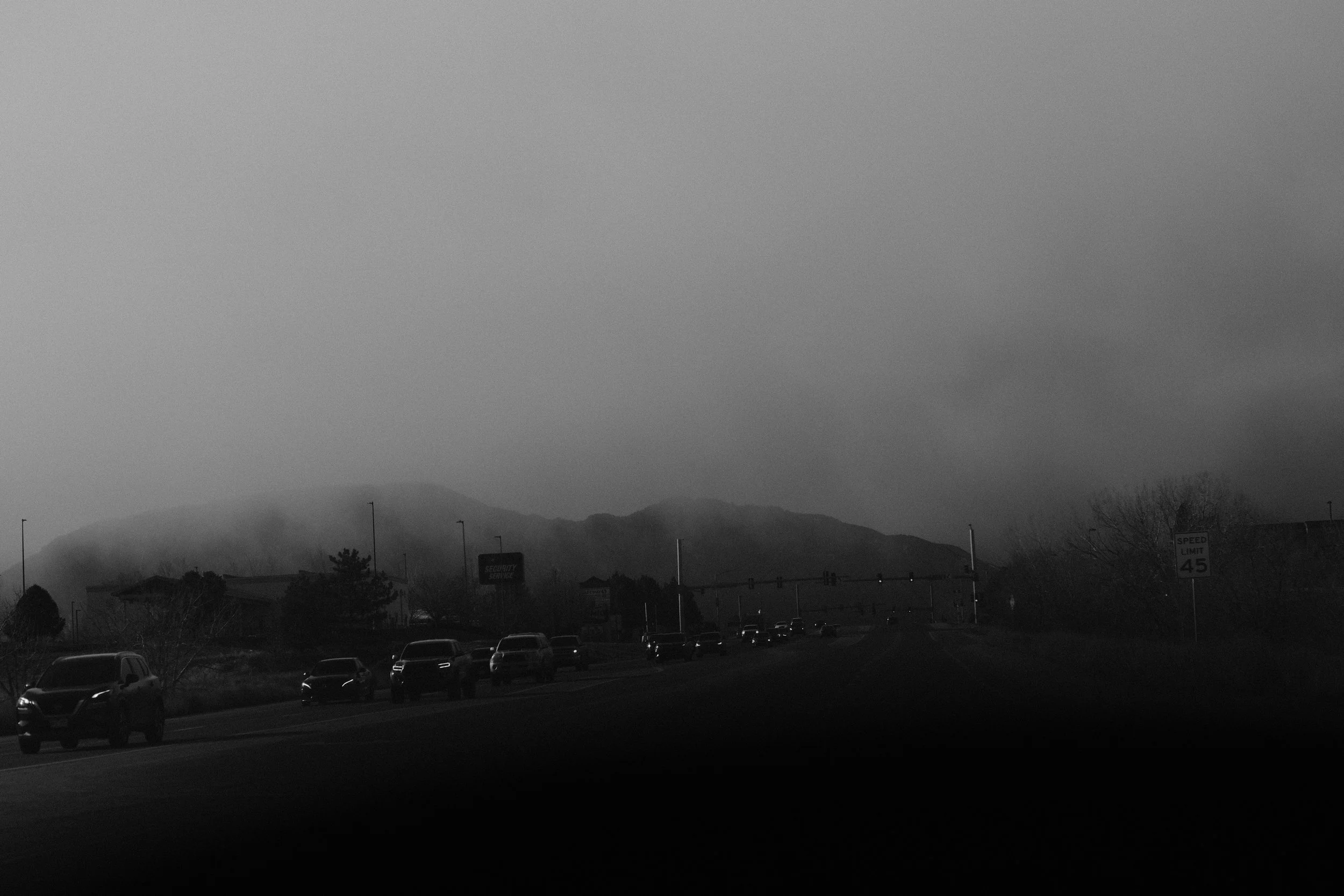 A grayscale photograph of a road with cars, traffic lights, street signs, and a mountain in the background overcast sky.