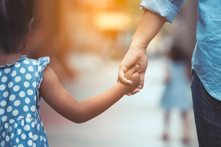 A young girl holding hands with an adult, possibly a parent, outdoors in the evening.
