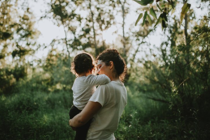 A woman holding a young child outdoors in a green, wooded area during sunlight.