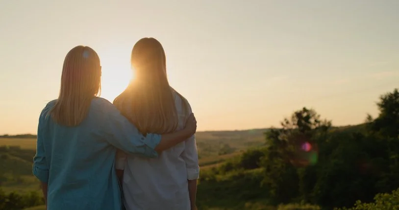 Two people with long hair standing outdoors at sunset, embracing, with a landscape of fields and trees in the background.
