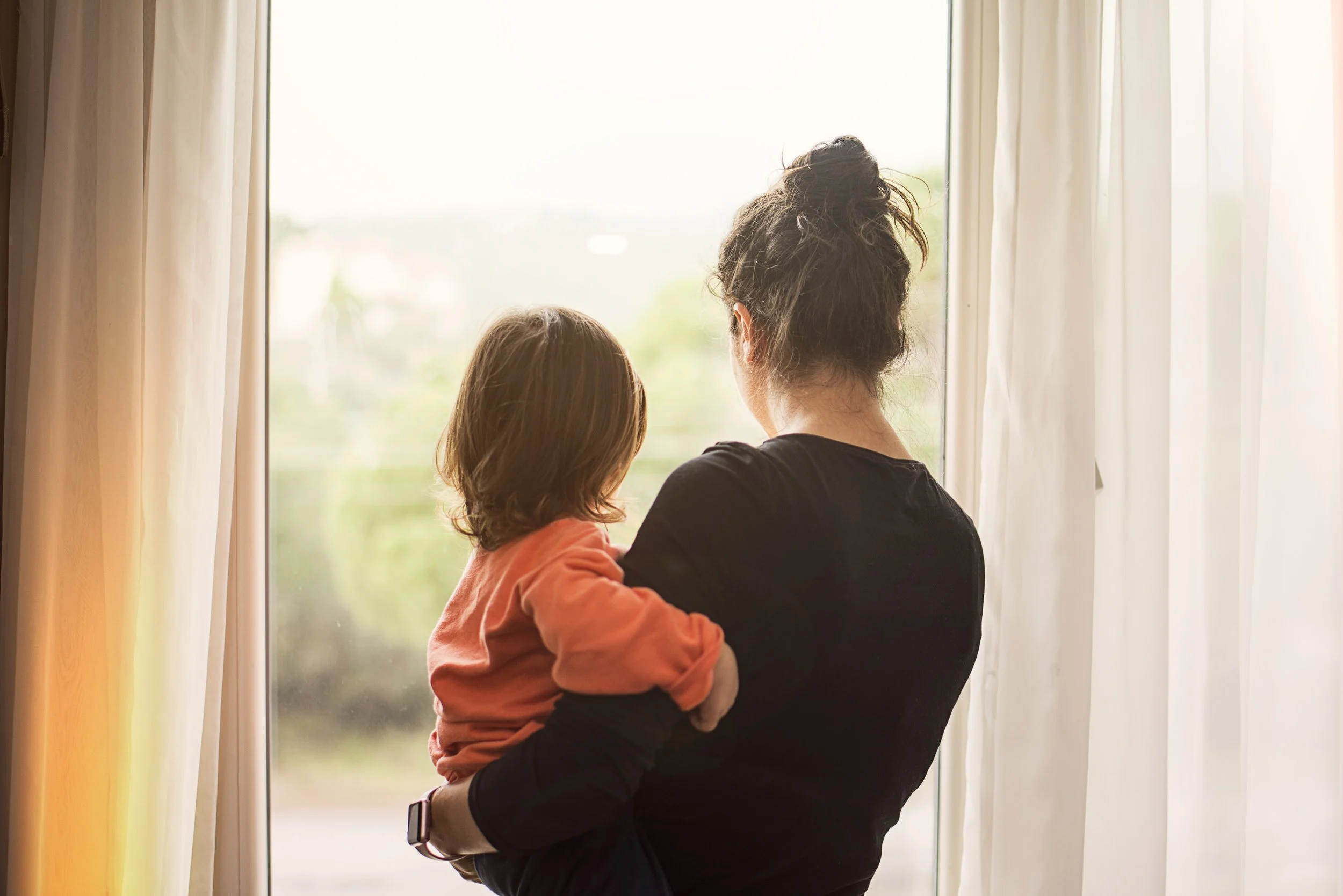A woman holding a young girl while looking out a large window with sheer curtains, with trees visible outside.