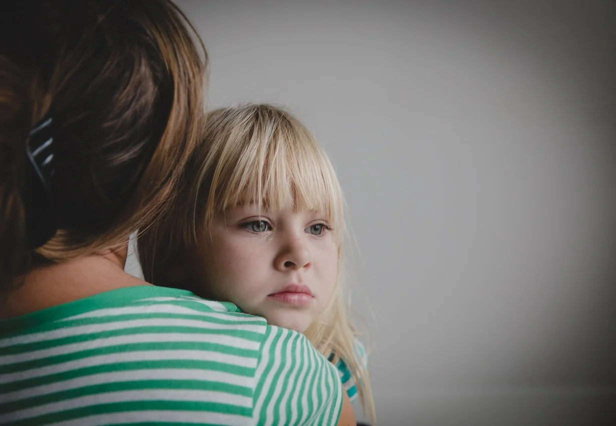 A woman holding a young girl with blonde hair and blue eyes, looking away with a serious expression, against a plain gray background.
