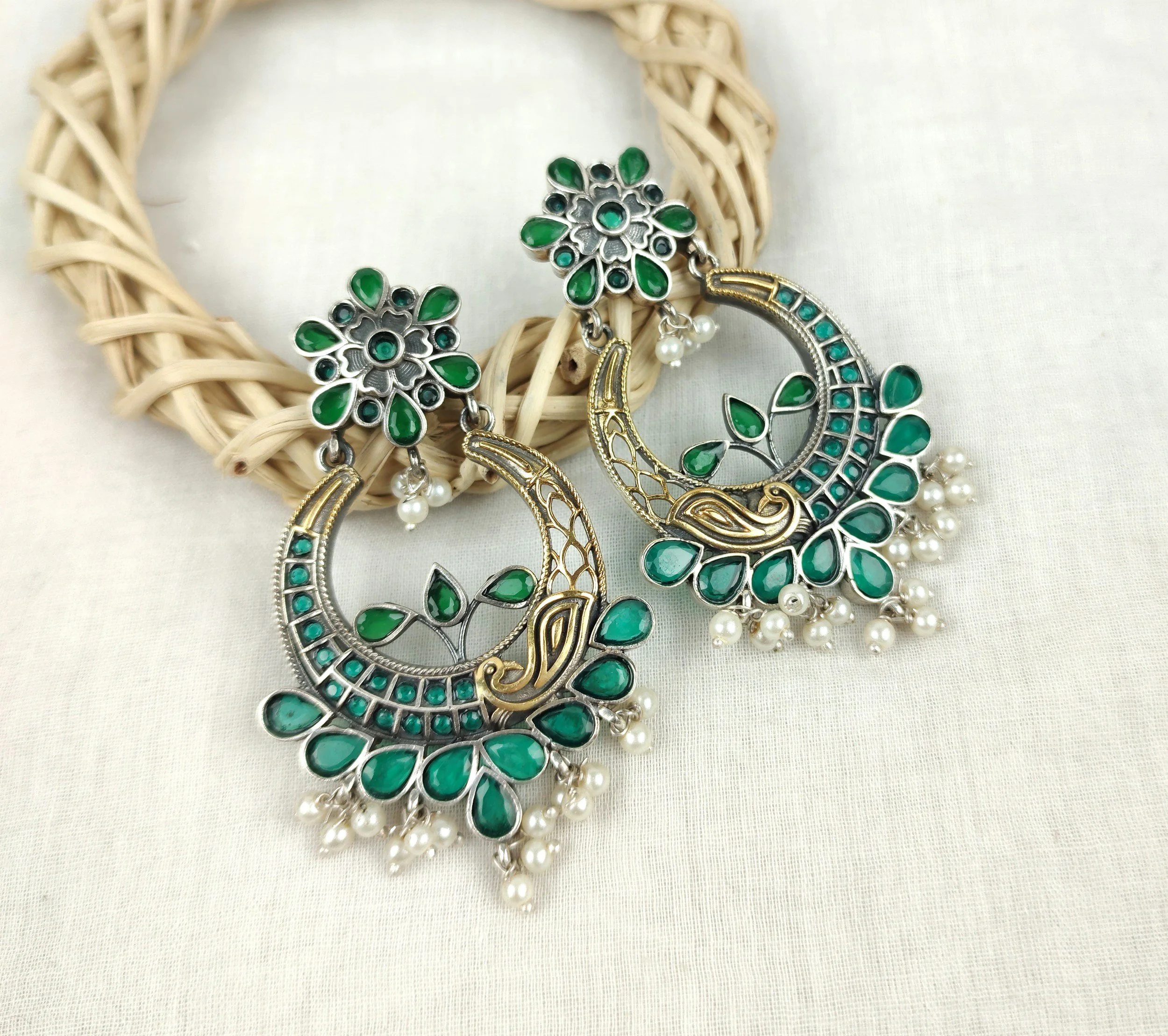 Pair of green and silver earrings with pearl accents on a white cloth background, positioned near a woven circular display.