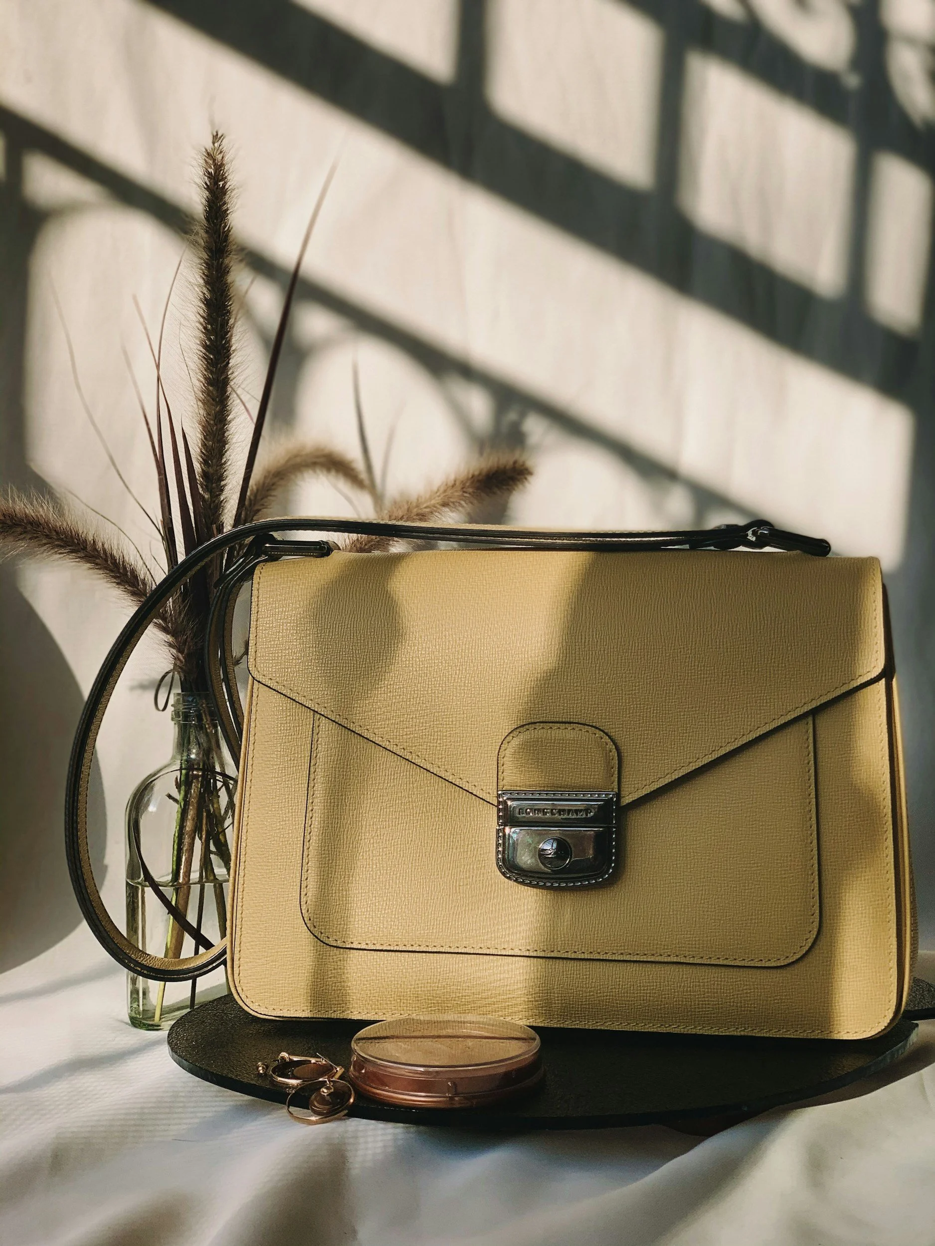 A yellow handbag with a silver lock, placed on a round black mat with a small round purse and a pair of earrings in front, next to a glass bottle with dried ornamental grass, with shadows cast on the wall behind.