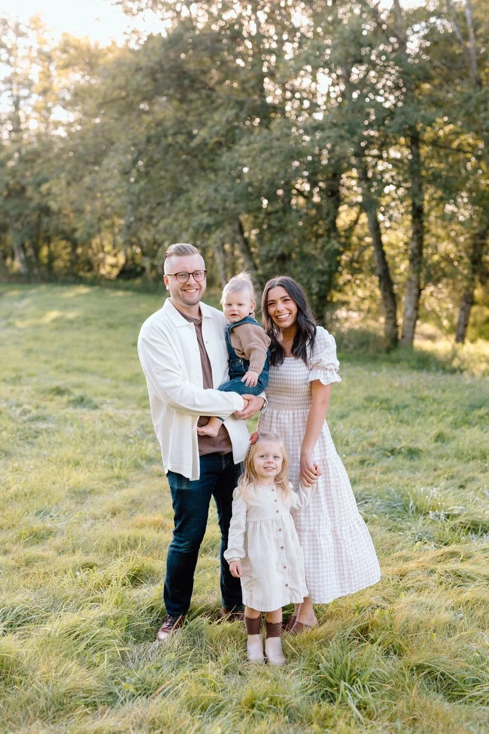 Family of four standing in a grassy field with trees in the background during golden hour, smiling at the camera.