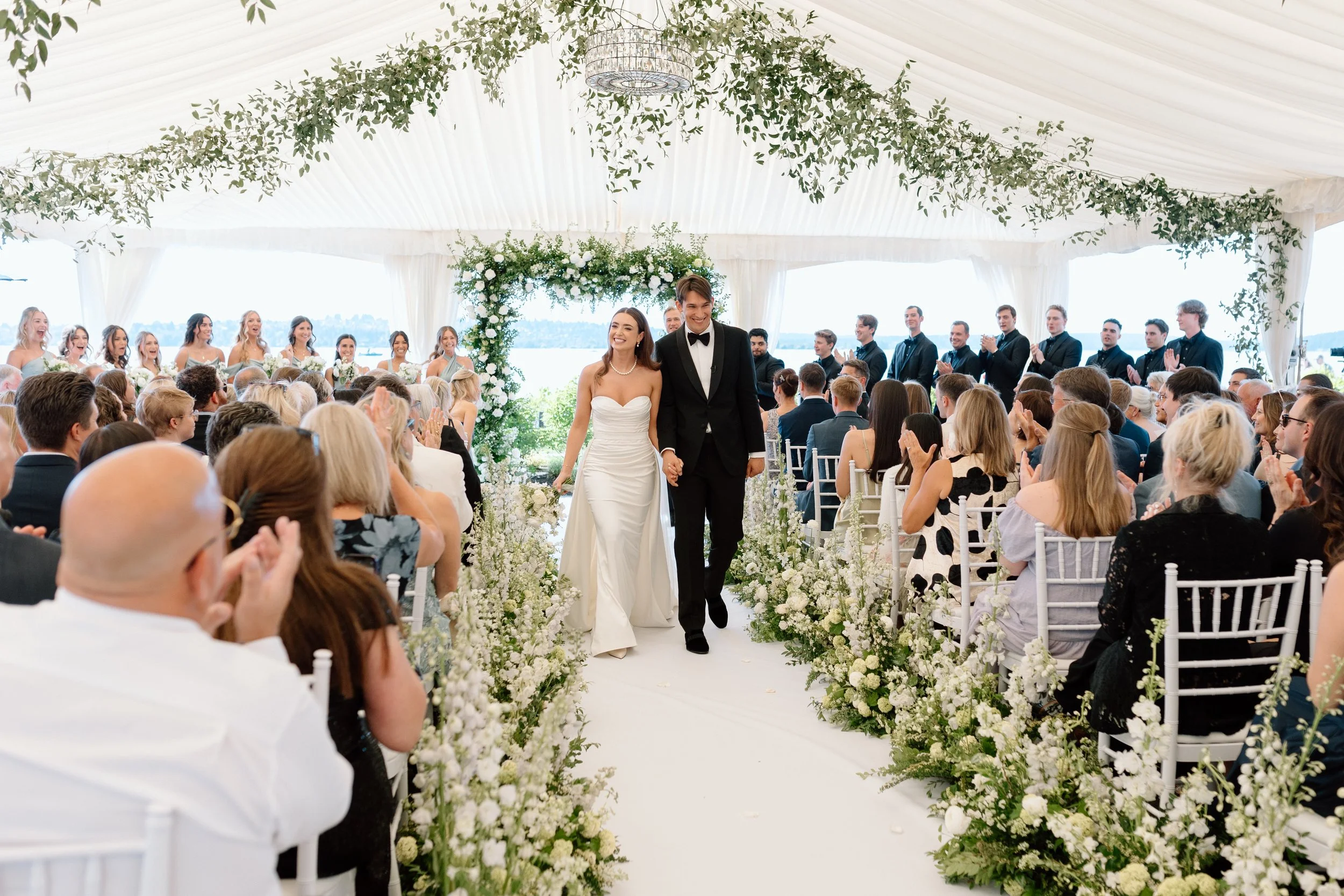 A bride and groom walking down the aisle at their outdoor wedding ceremony, surrounded by white flowers and greenery, with guests clapping and smiling.