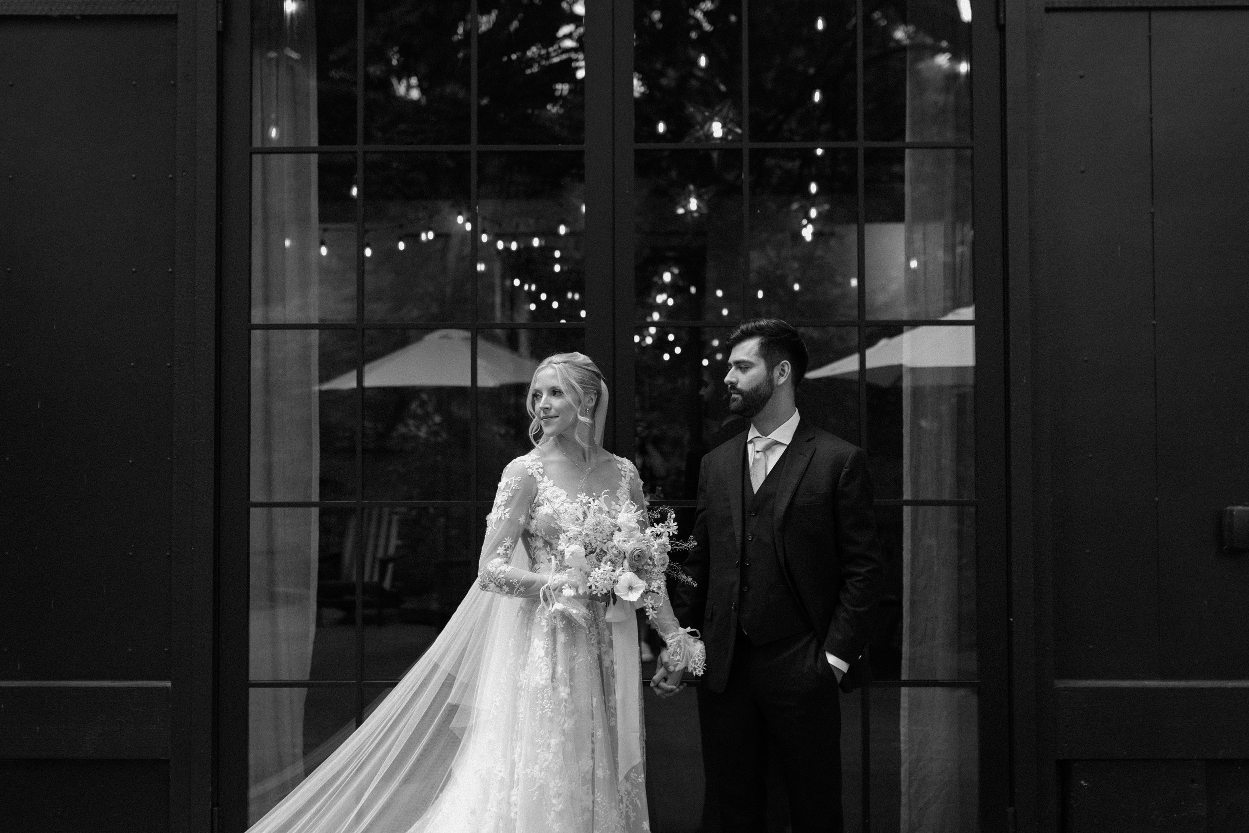 A newlywed couple stands hand in hand inside a building with large glass windows, reflecting string lights outside. The bride wears a lace wedding dress with a long train and holds a bouquet of flowers, while the groom wears a dark suit with a vest and tie. The scene is black and white, with a romantic and elegant atmosphere.