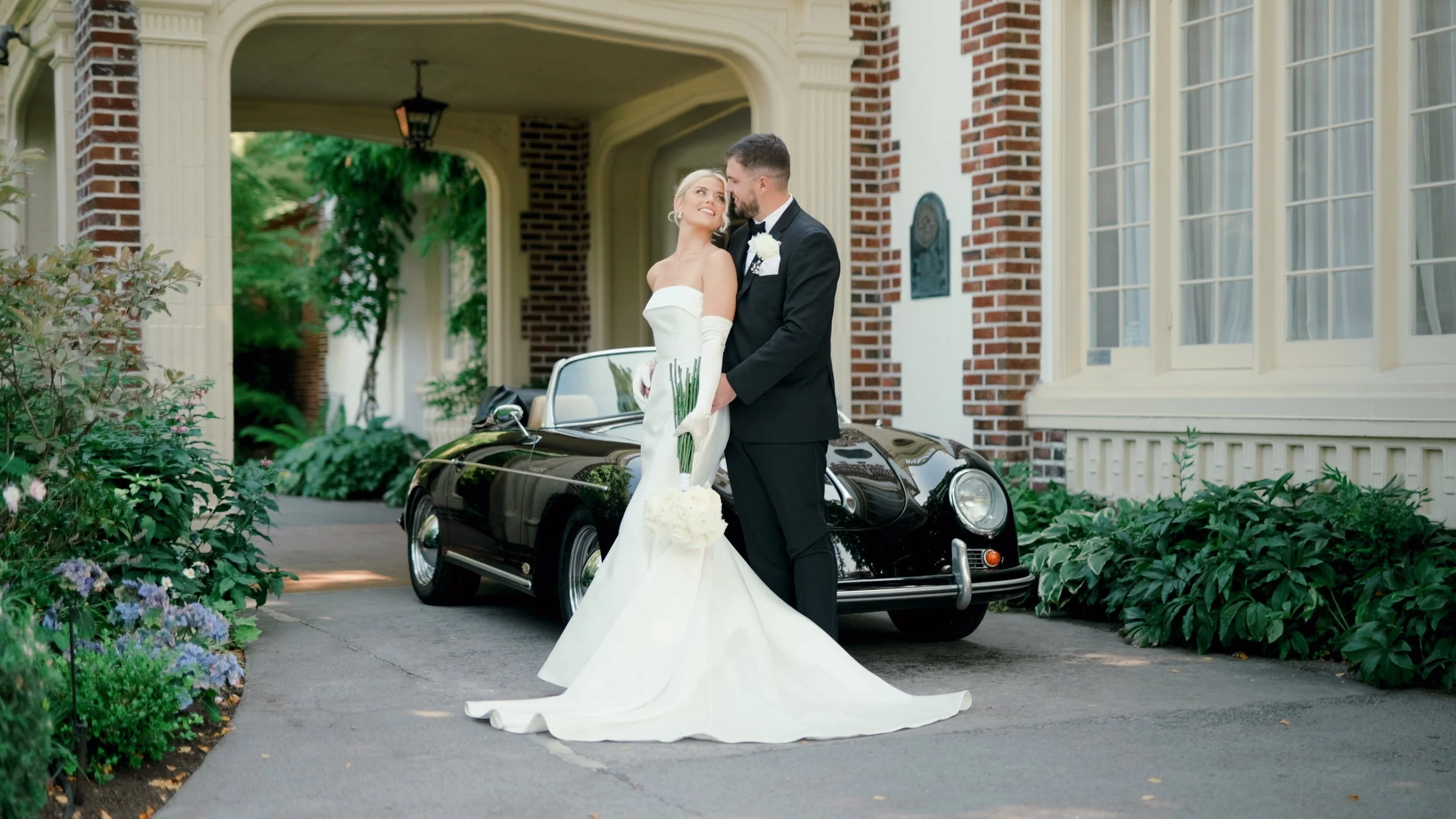 A bride and groom standing next to a vintage black car in front of a brick and cream-colored house, surrounded by greenery and flowers, during a wedding day.