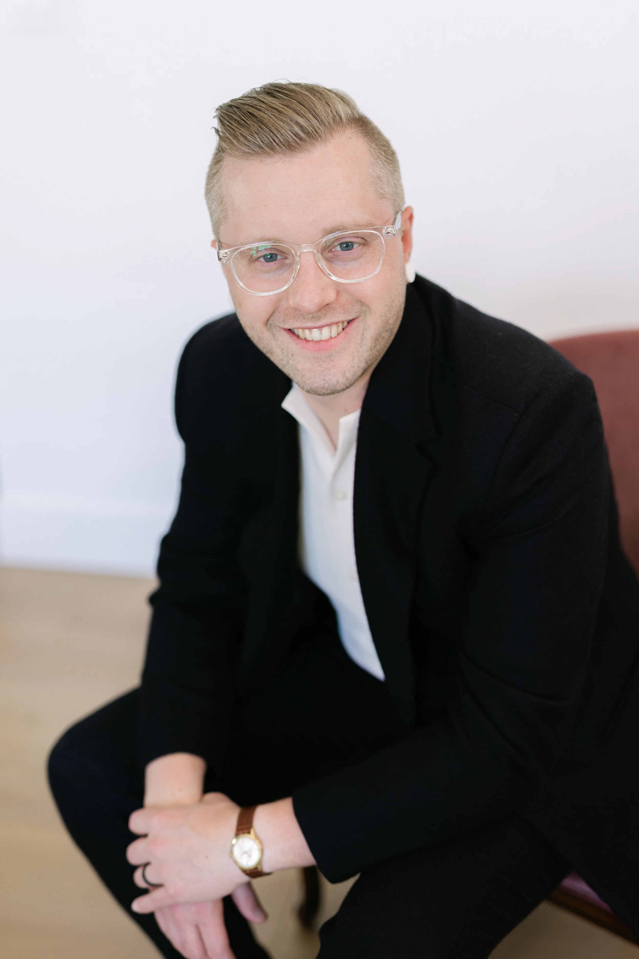 A young man with blonde hair, wearing clear glasses, a black blazer, and white shirt, smiling and seated with hands clasped, against a white background.