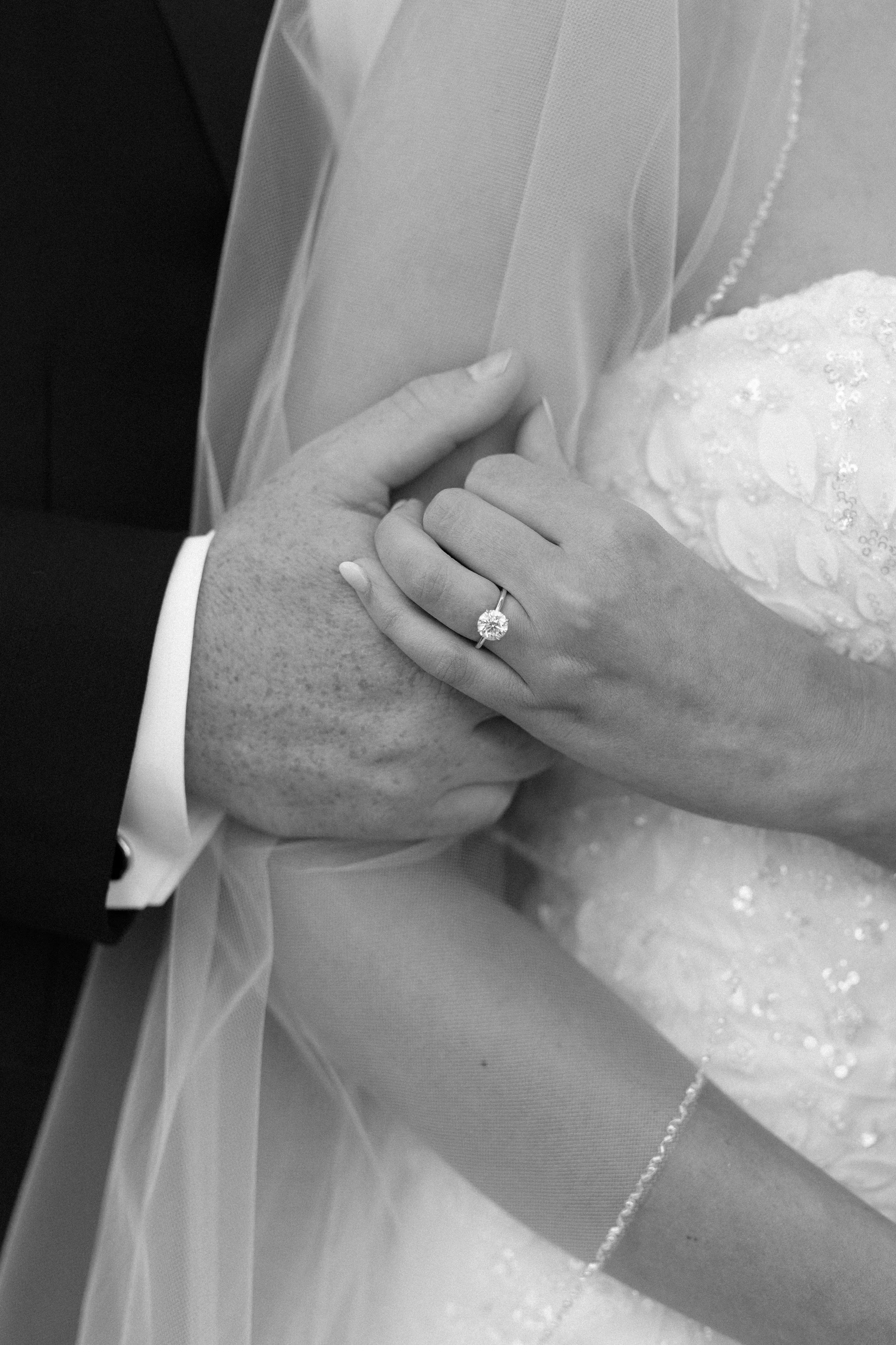 A close-up of a bride and groom holding hands, showing an engagement ring and wedding band on her finger, with a bridal dress and veil.