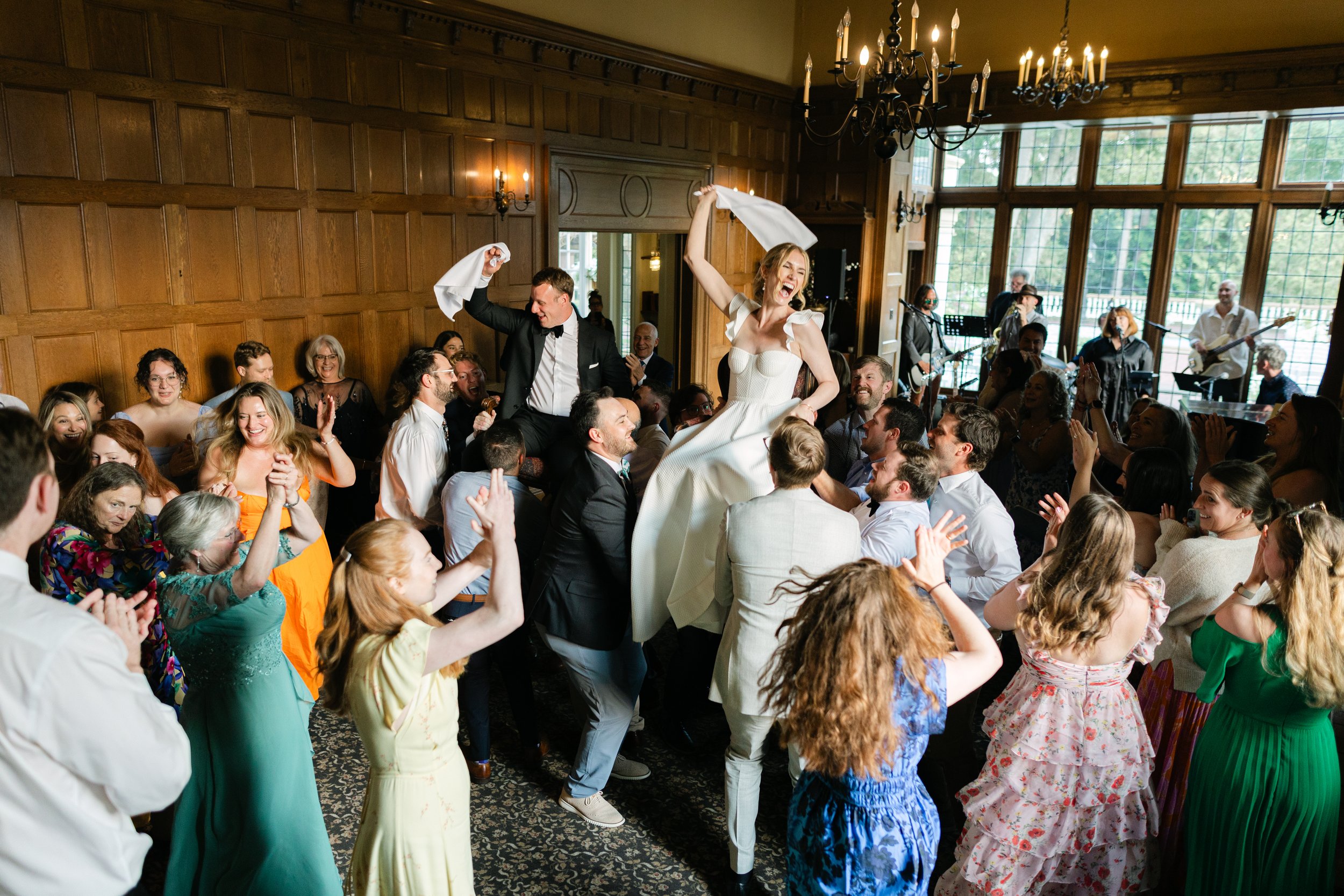 Wedding reception with a dance floor where guests are celebrating and dancing. The bride and groom are being lifted on chairs, dancing, with a live band playing in the background. The room has wooden-paneled walls, chandeliers, and large windows letting in natural light.