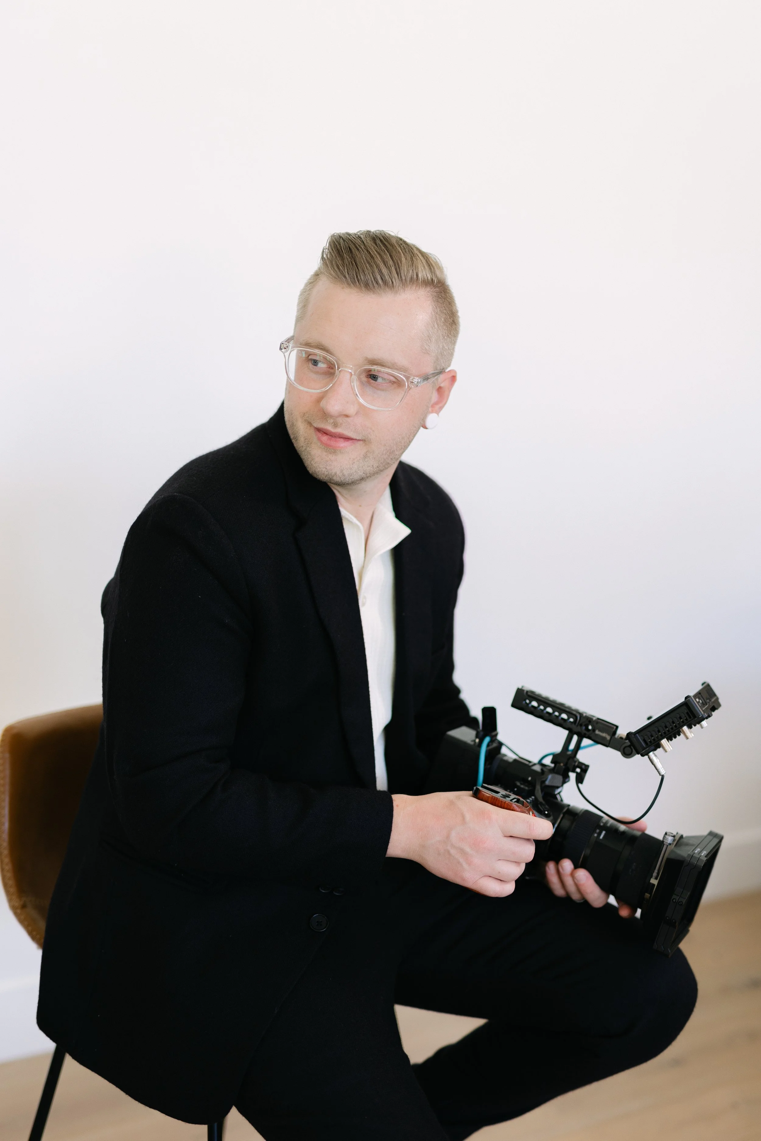 A man with blonde hair, wearing glasses, a white shirt, and a black blazer, seated on a chair against a white wall, operating a camera stabilizer equipment.