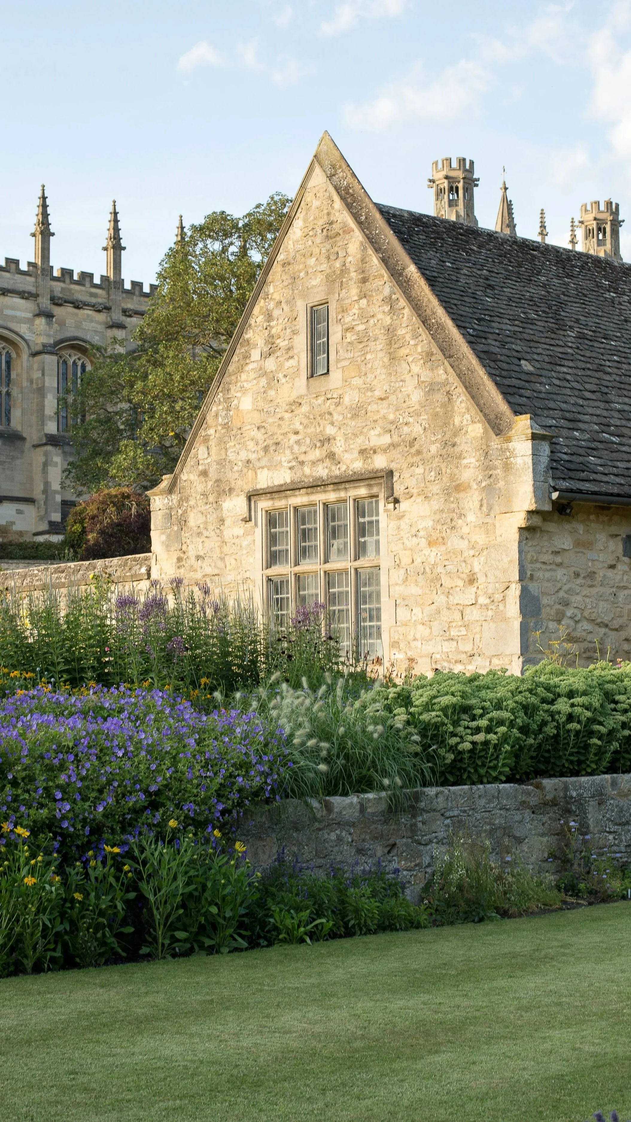 A stone house with a steep roof, multiple chimneys, and a large window, surrounded by a garden with purple and yellow flowers, green plants, and a well-manicured lawn, with a church in the background.
